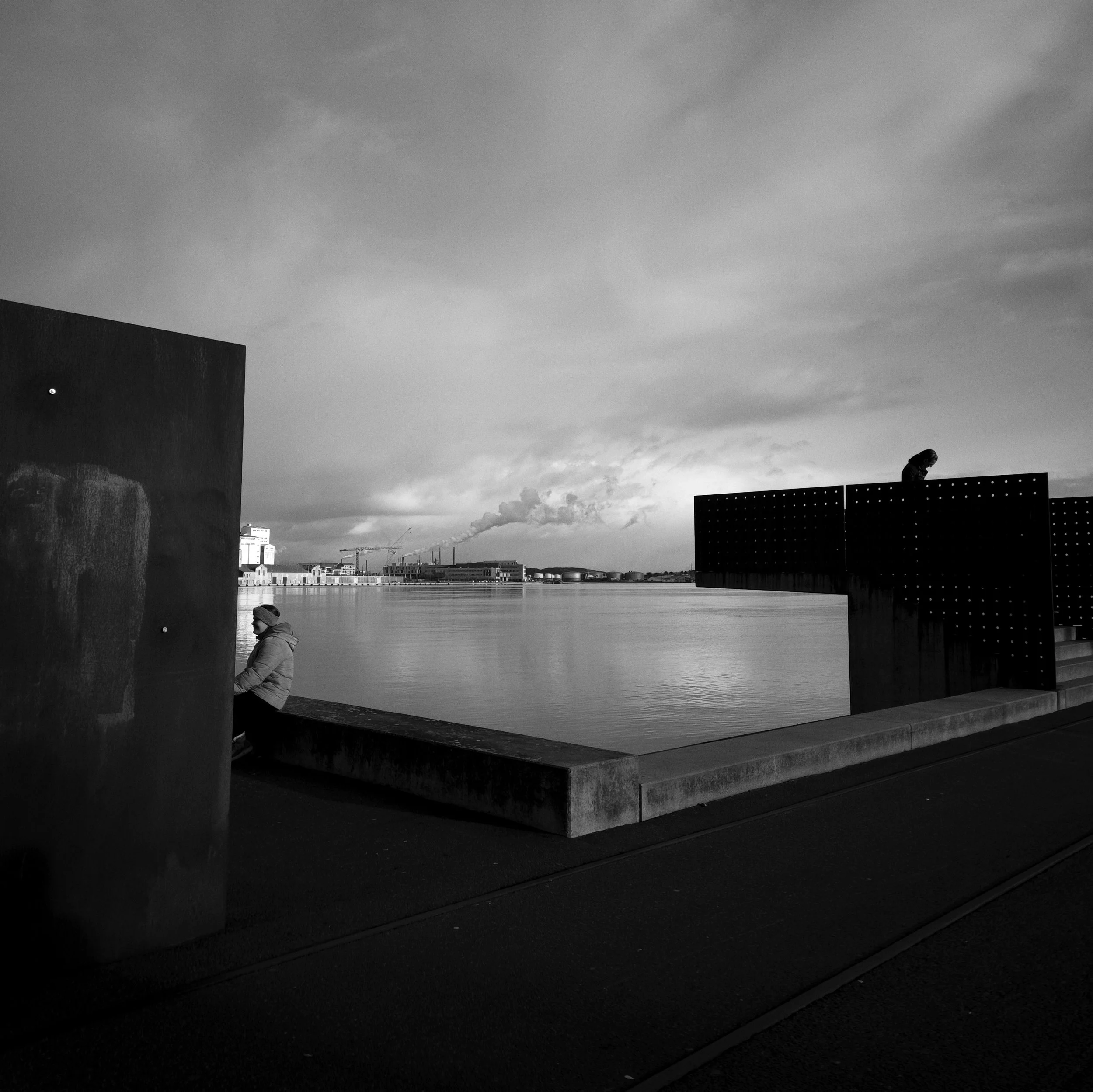 Black and white photo of a waterfront promenade with two people, one sitting on the edge and another walking on the path, with industrial buildings and smokestacks in the background.