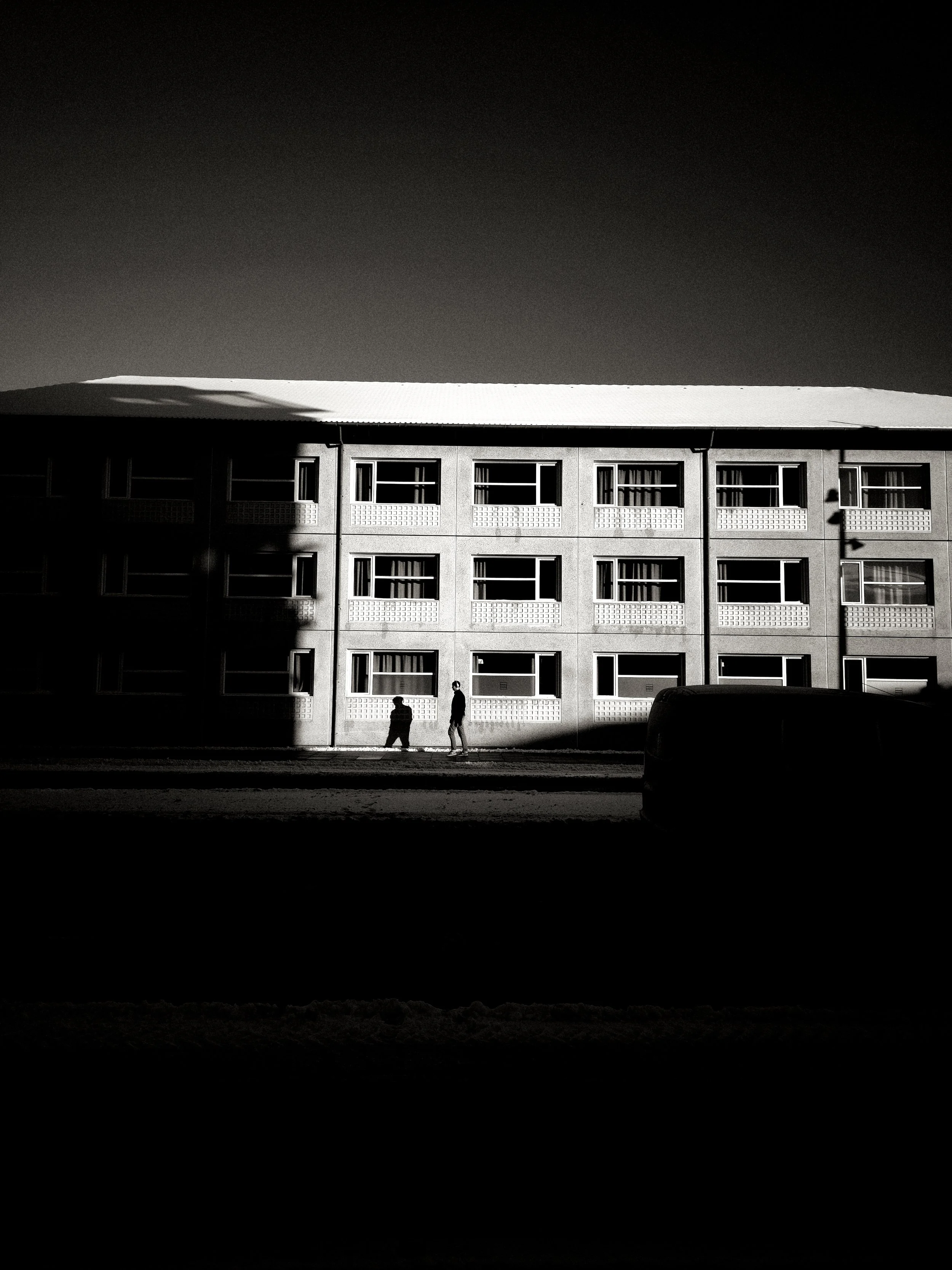 Black and white photo of a building with multiple windows, shadows cast on the lower part of the building, and two children walking at the sidewalk, with a car partially visible in the foreground.