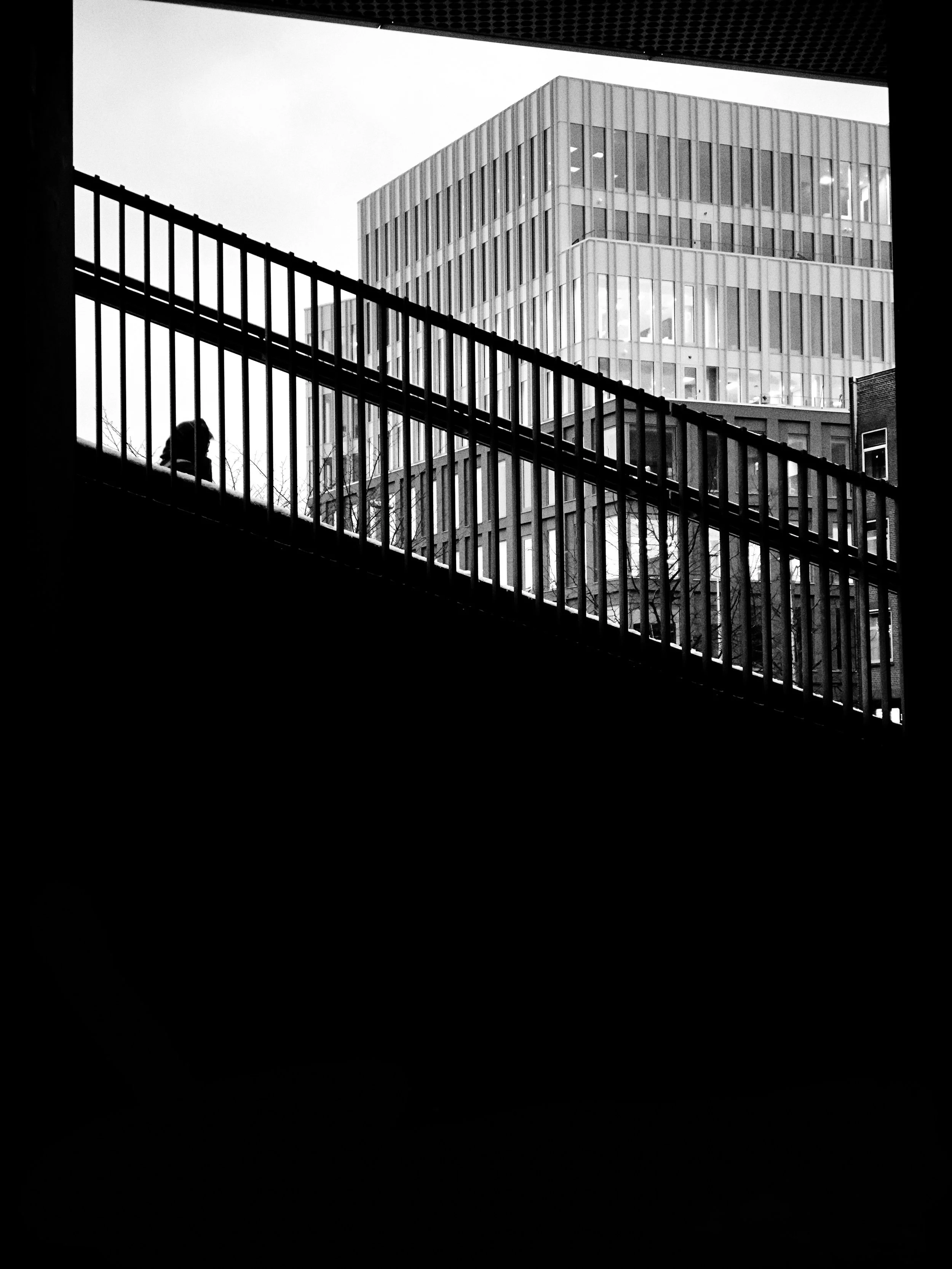 Black and white photo of a person walking on a bridge with vertical bars, with modern multi-story buildings in the background.