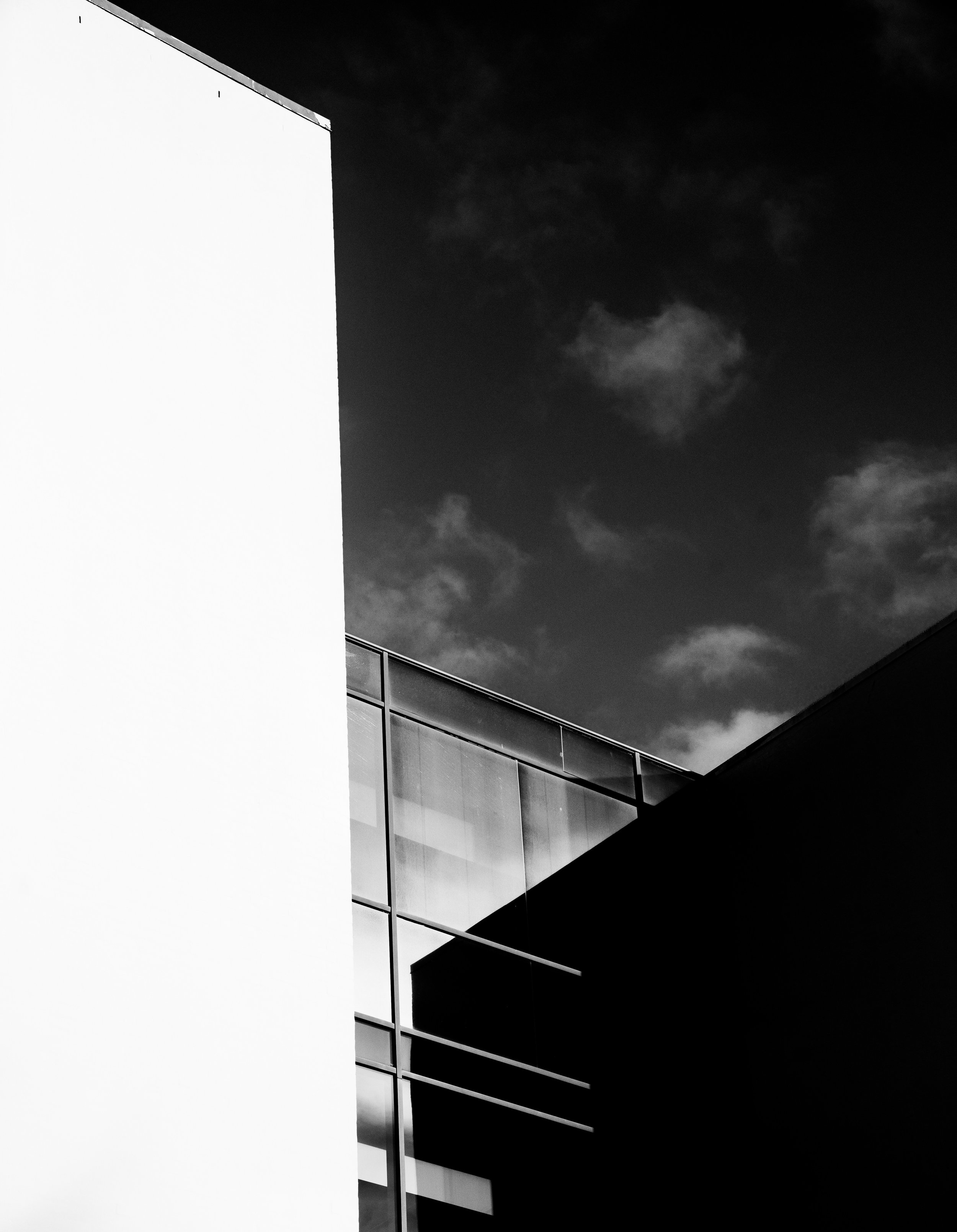 Black and white photo of modern building with sharp lines, white wall on the left, glass windows in the middle, and a dark section on the right, with a cloudy sky above.