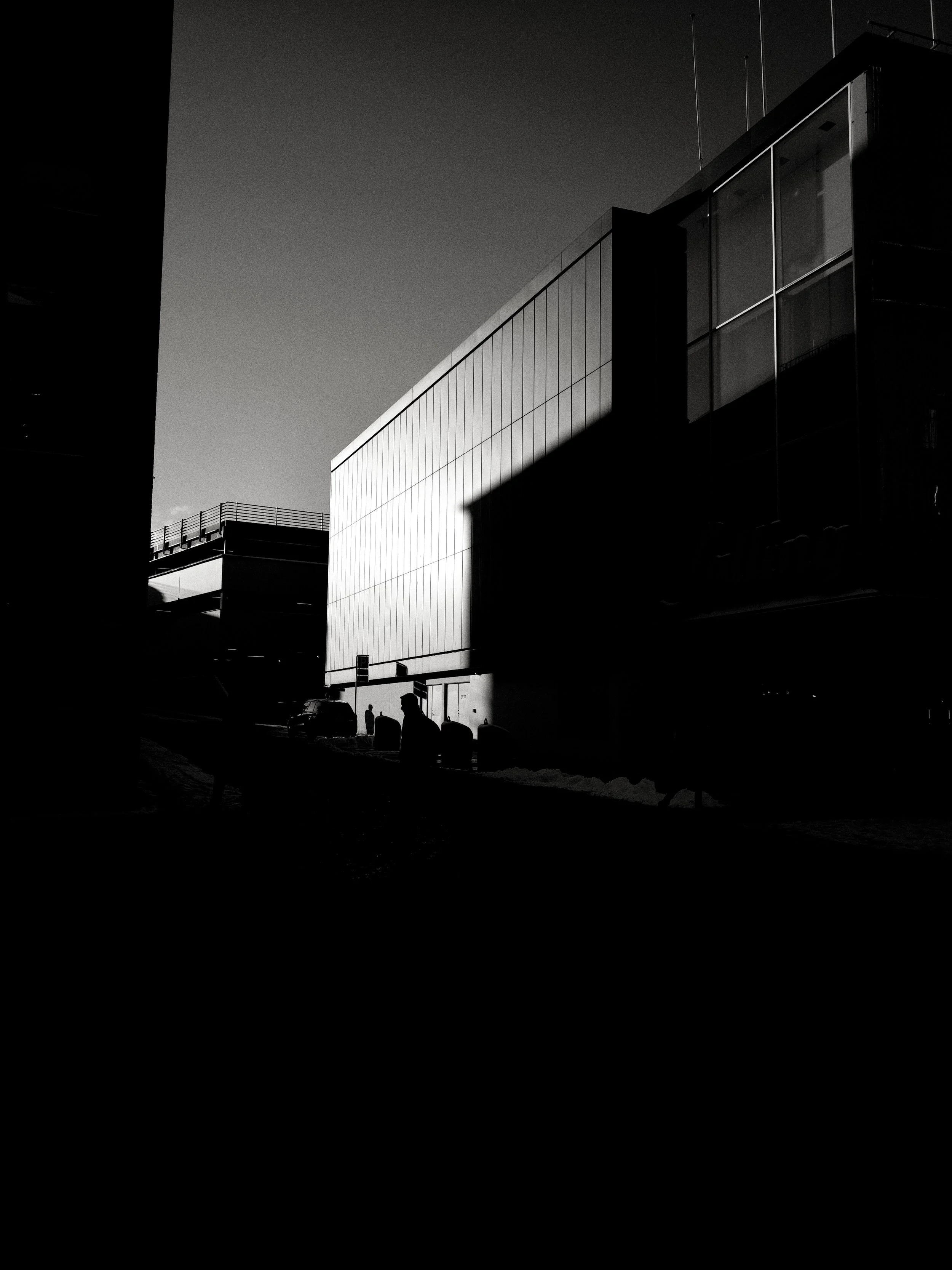 Black and white photo of urban architecture with modern buildings, silhouettes of people, and parked cars, with long shadows indicating late afternoon or early evening.