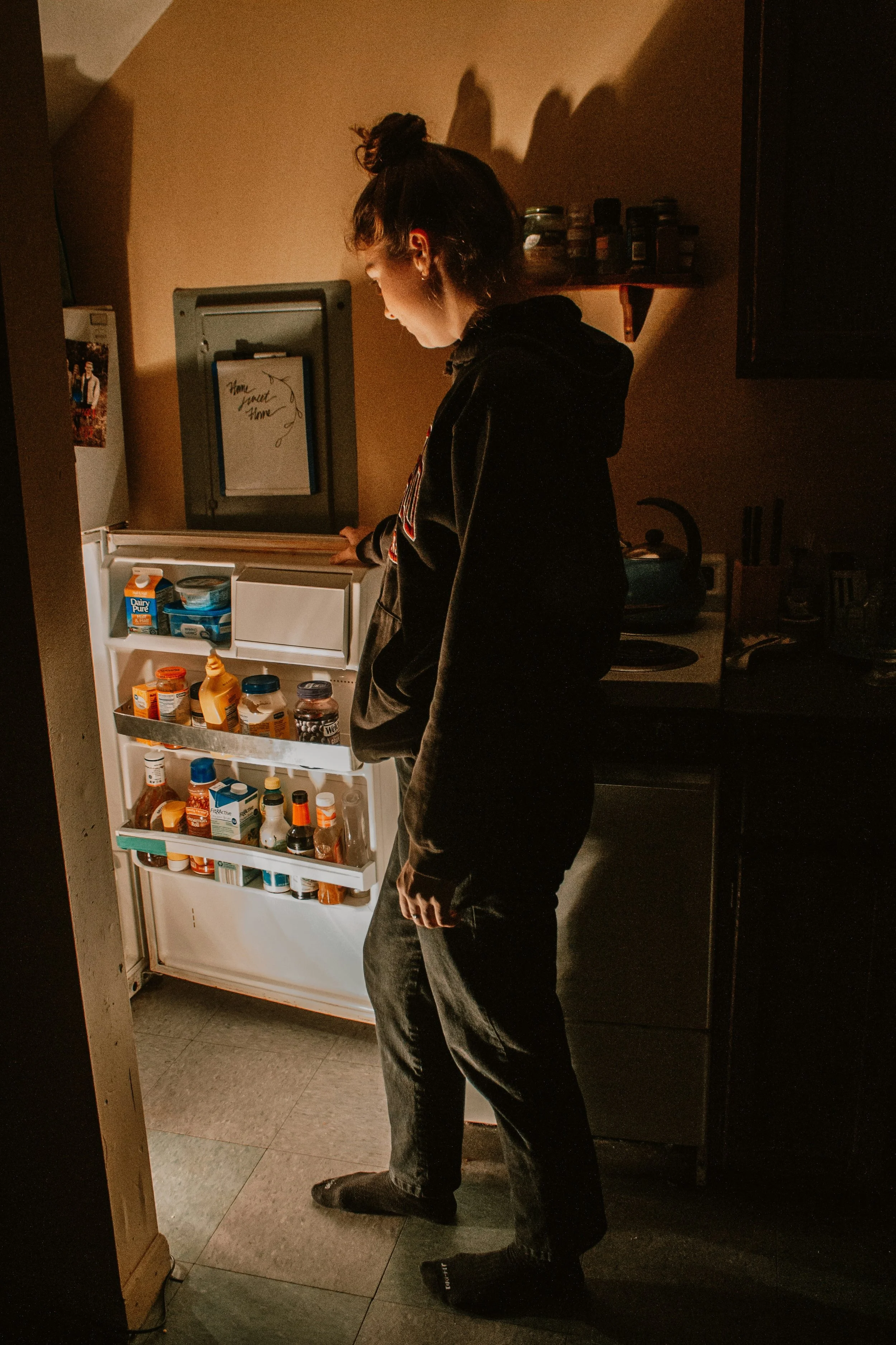 auburn haired woman looking in the open refrigerator pensively at night