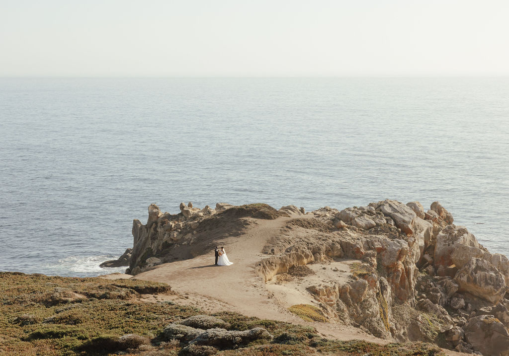 Bride in white gown and groom walking through gold grass field near Sea Ranch ocean cliffs