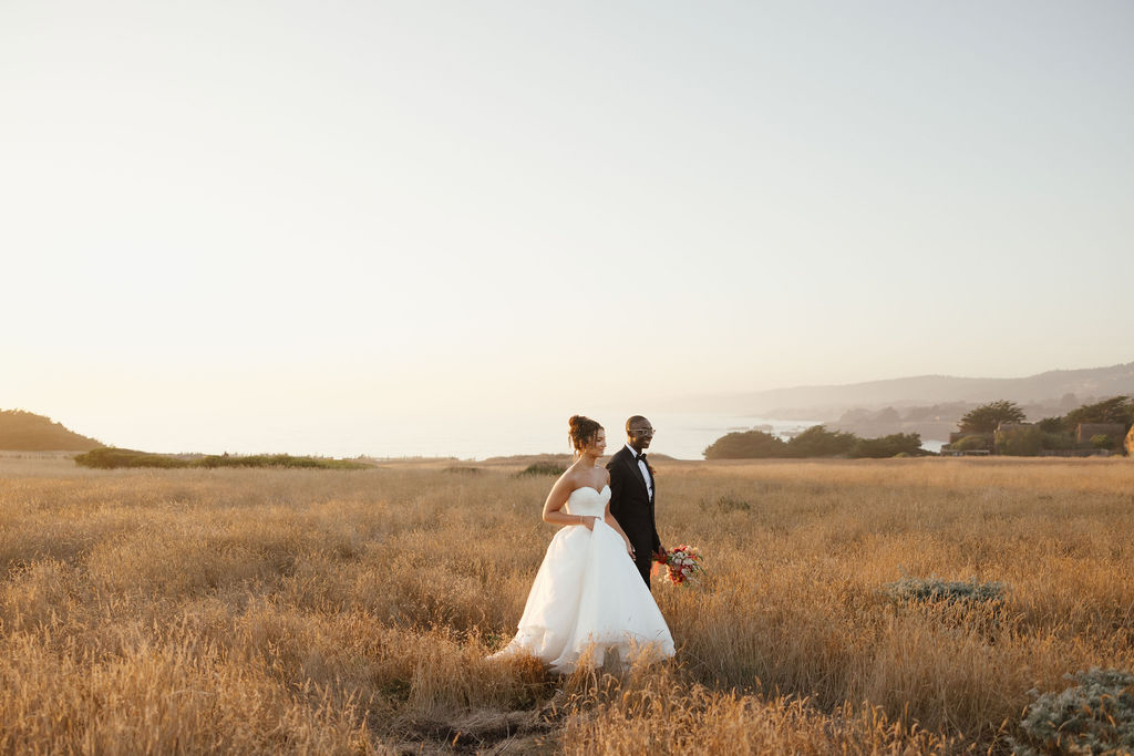Bride in white gown and groom walking through gold grass field near Sea Ranch ocean cliffs