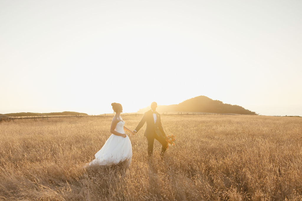 Bride in white gown and groom walking through gold grass field near Sea Ranch ocean cliffs