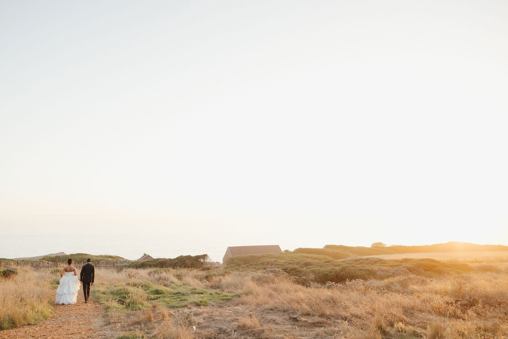 Bride in white gown and groom walking through gold grass field near Sea Ranch ocean cliffs