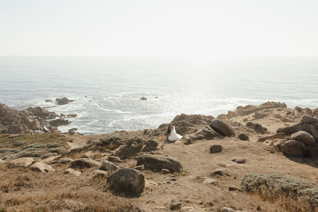 Bride in white gown and groom walking through gold grass field near Sea Ranch ocean cliffs