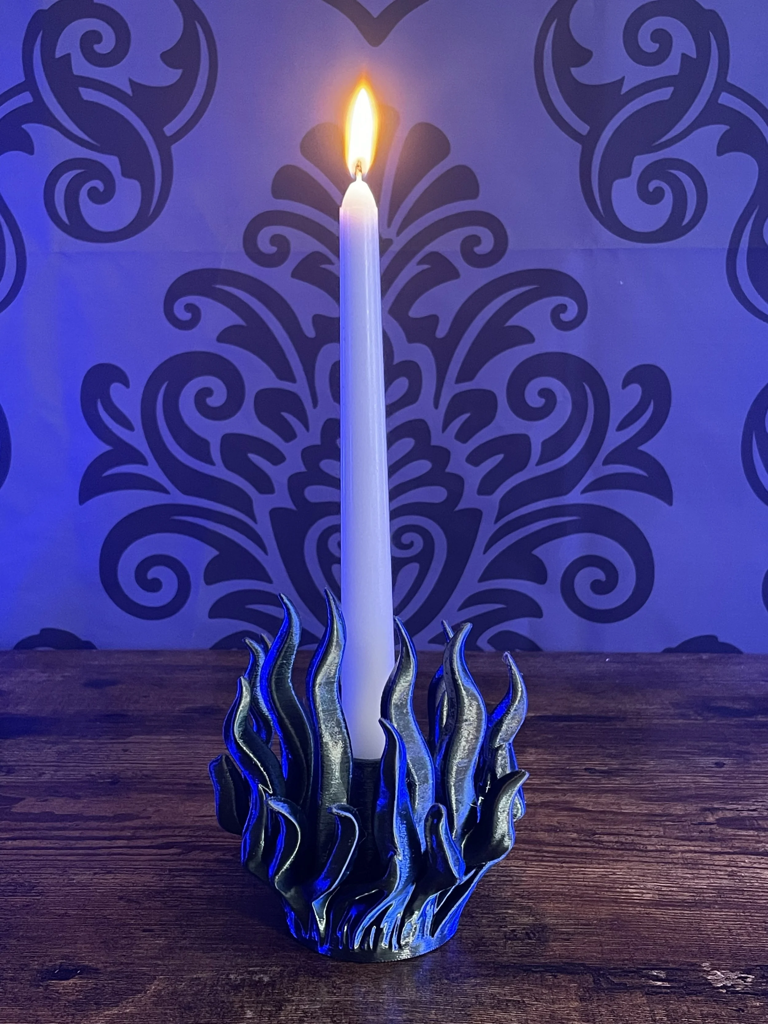 A lit white candle placed in a decorative black and gold glass holder with flame glowing, set on a wooden surface with a dark blue patterned background.