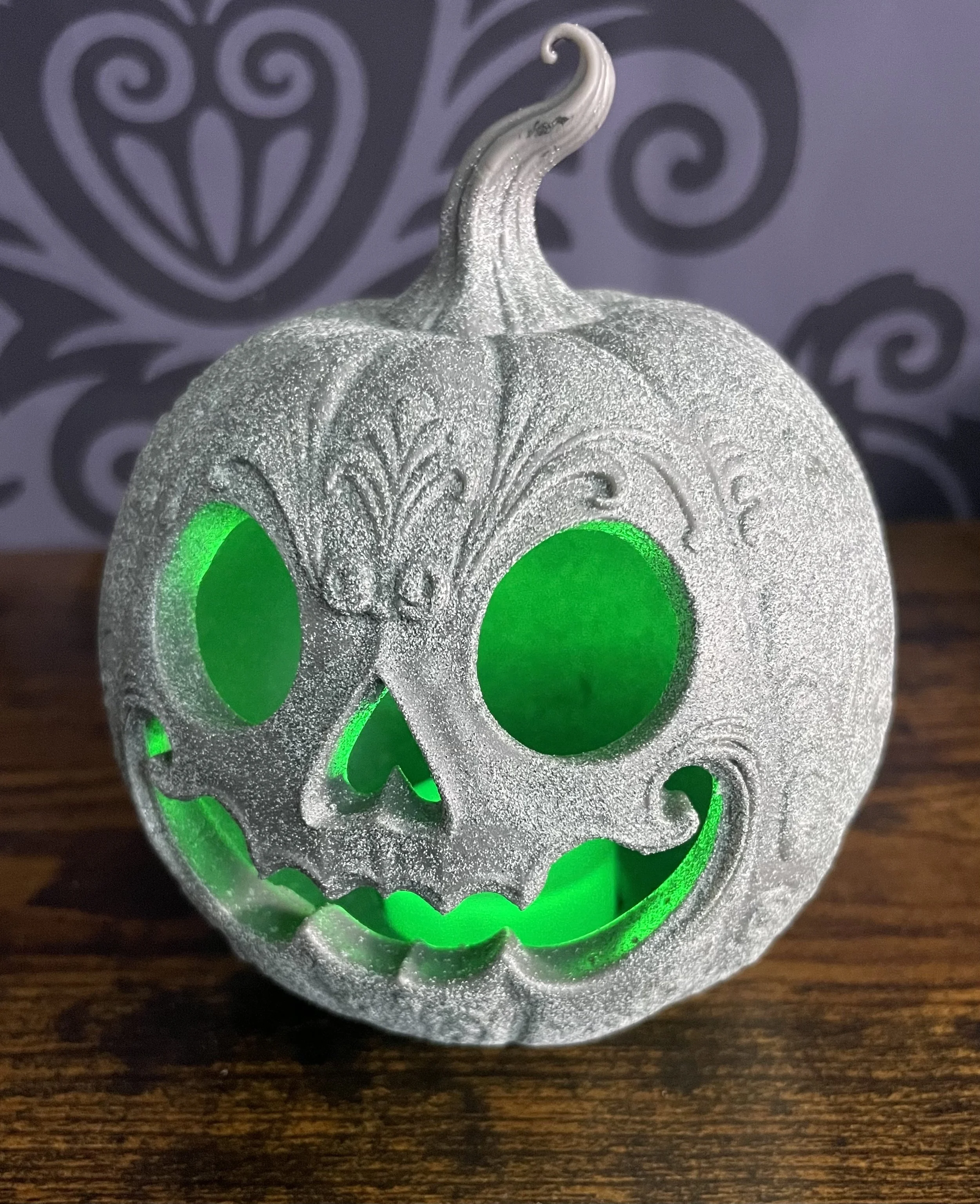 A decorative pumpkin with a glowing green skull face carved into it, with detailed engravings and a glittery silver finish, placed on a wooden surface against a dark background with swirling patterns.