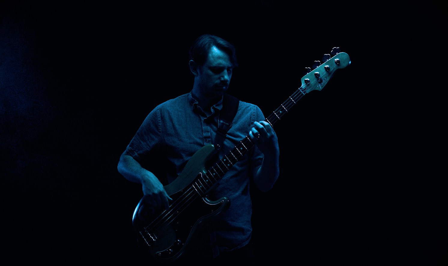 A man with shoulder-length brown hair and a beard, wearing a red shirt, sits on a dark red velvet couch in front of a black background with red vertical lines, illuminated by blue and red lighting.