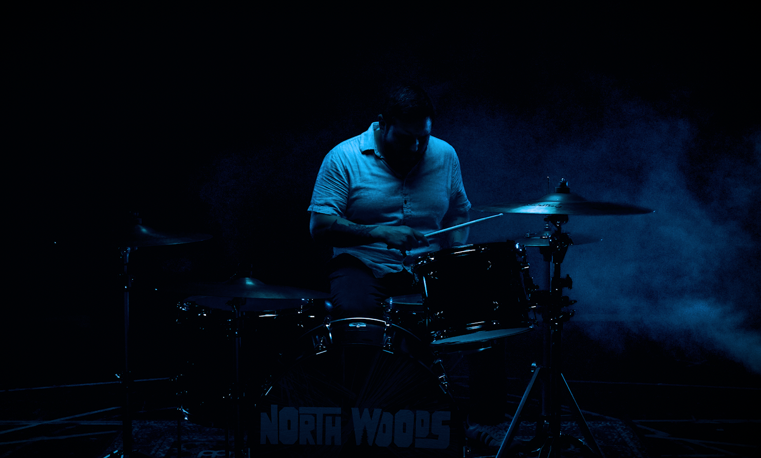 A man with tattoos on his arm, sitting at a table with his hands clasped, looking directly at the camera in a dark room with red and blue lighting.