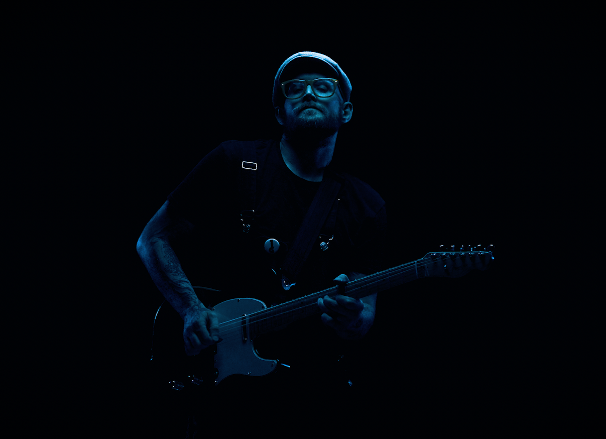 A man with glasses, a beanie, and a beard sitting on a red velvet couch with red curtains in the background, illuminated by red and teal lighting.