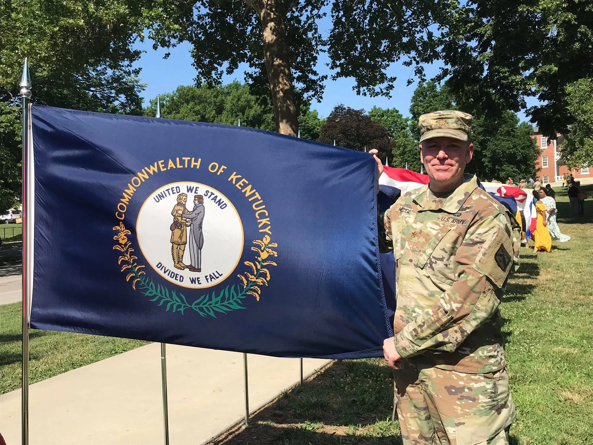 Jonathan Holliday in camouflage uniform holding a Kentucky state flag with the state seal, outdoors on a sunny day with trees and people in the background at his military graduation from US Army Command and General Staff College.