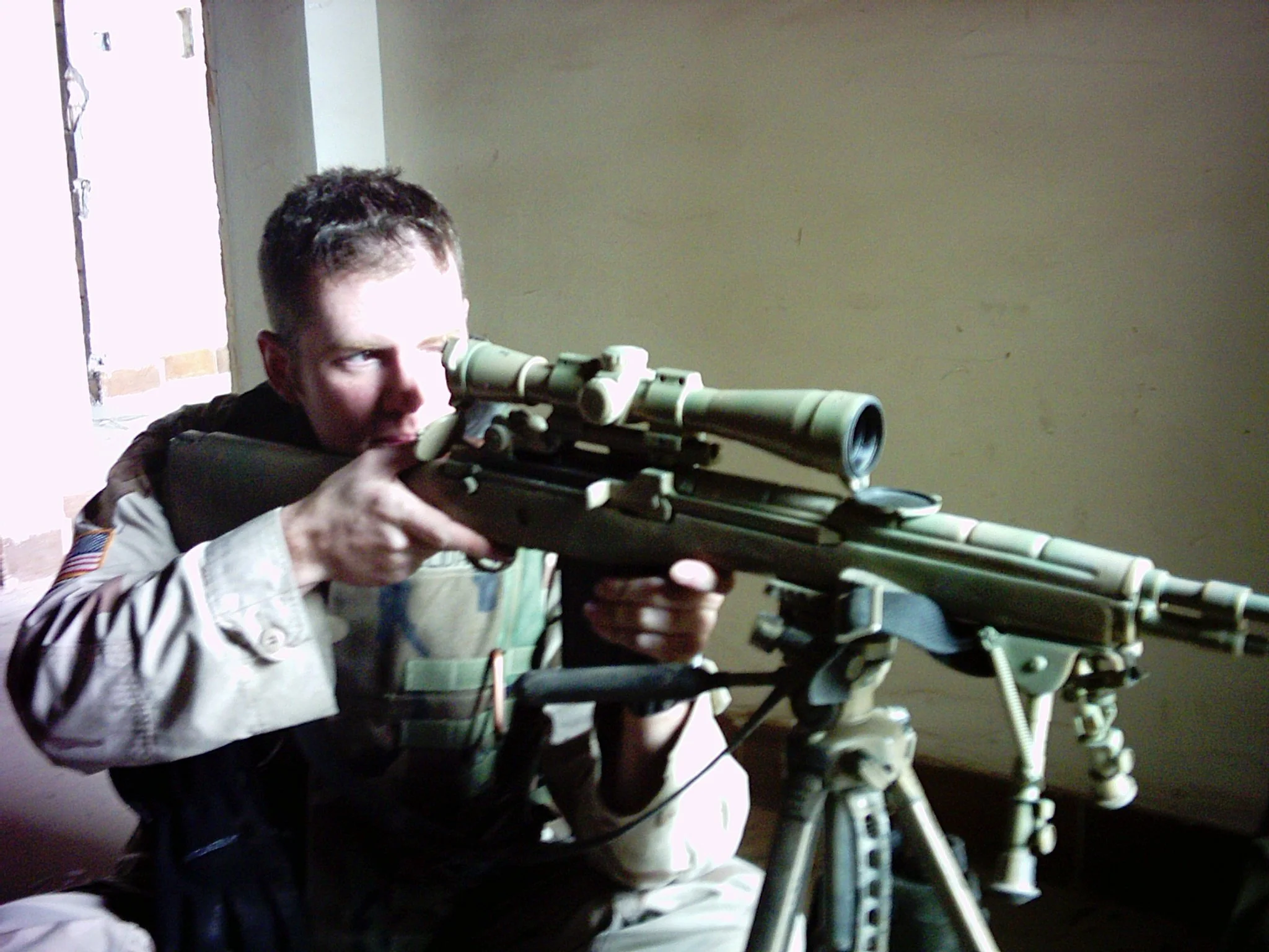 A young boy in military-style clothing aiming a large sniper rifle through a scope indoors.