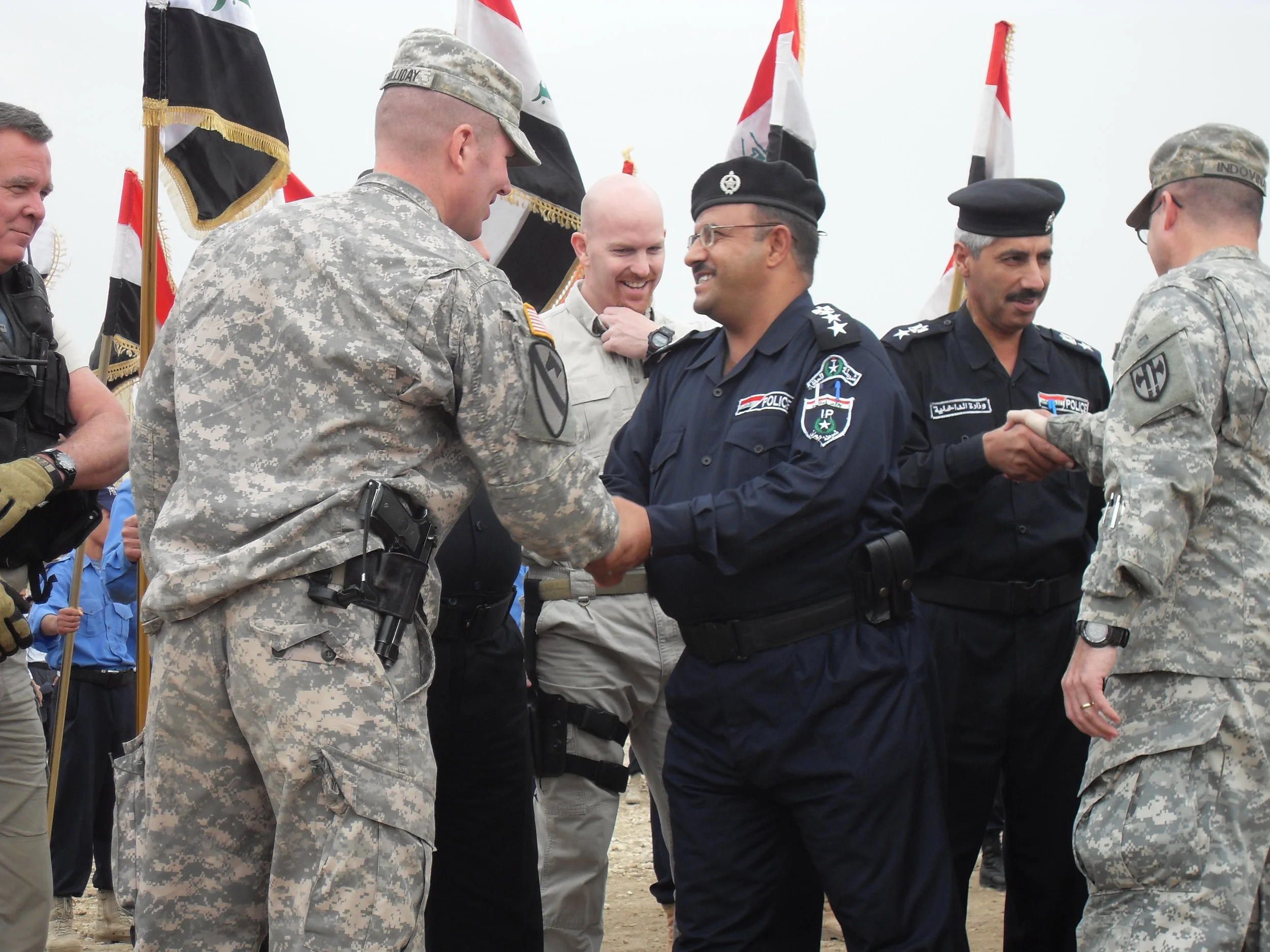 Jonathan Holliday, US military personnel and Iraqi police officers shaking hands at a ceremony, with flags in the background in Baghdad, Iraq.