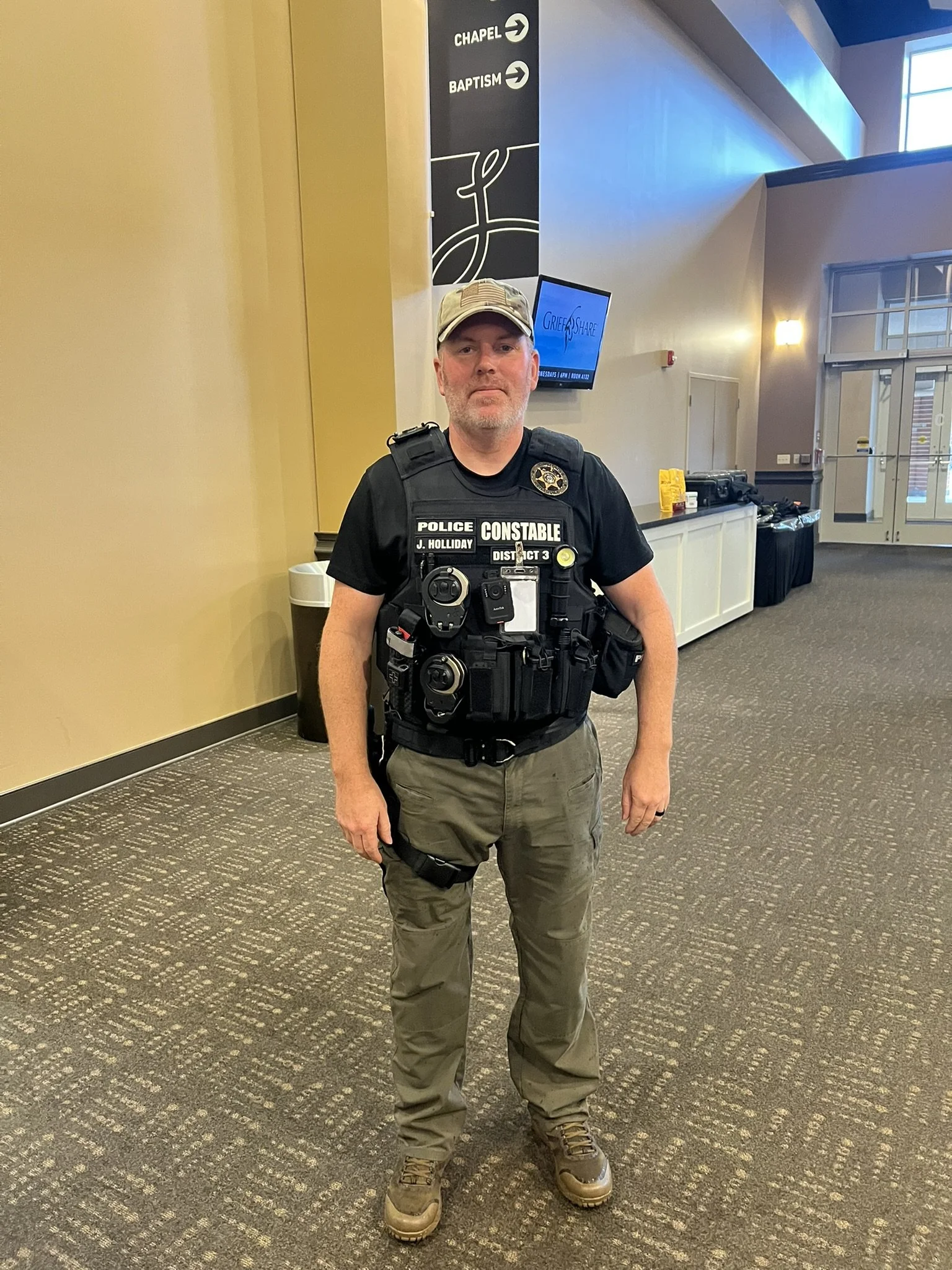 A police officer wearing tactical gear, including a bulletproof vest, stands inside a building with beige walls and carpeted floor. The vest has various patches and equipment attached. Behind him, there is a wall with directional signs pointing to th