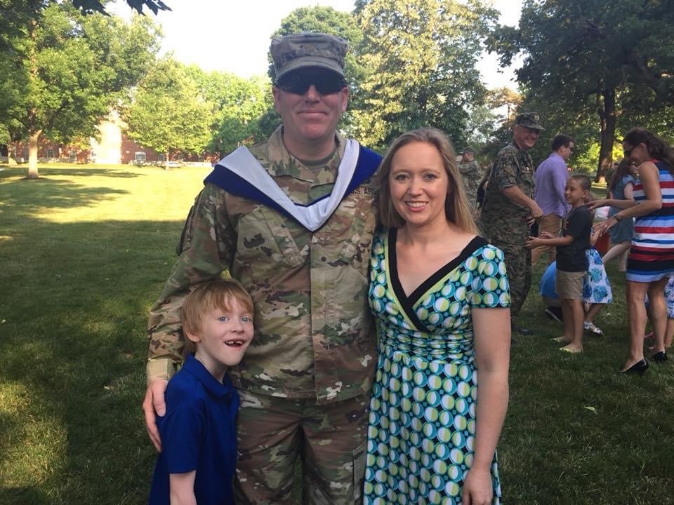 A soldier in camouflage uniform and sunglasses standing with a woman in a patterned dress and a young boy in a blue shirt outdoors in a park. Several people are in the background, some dancing or socializing.