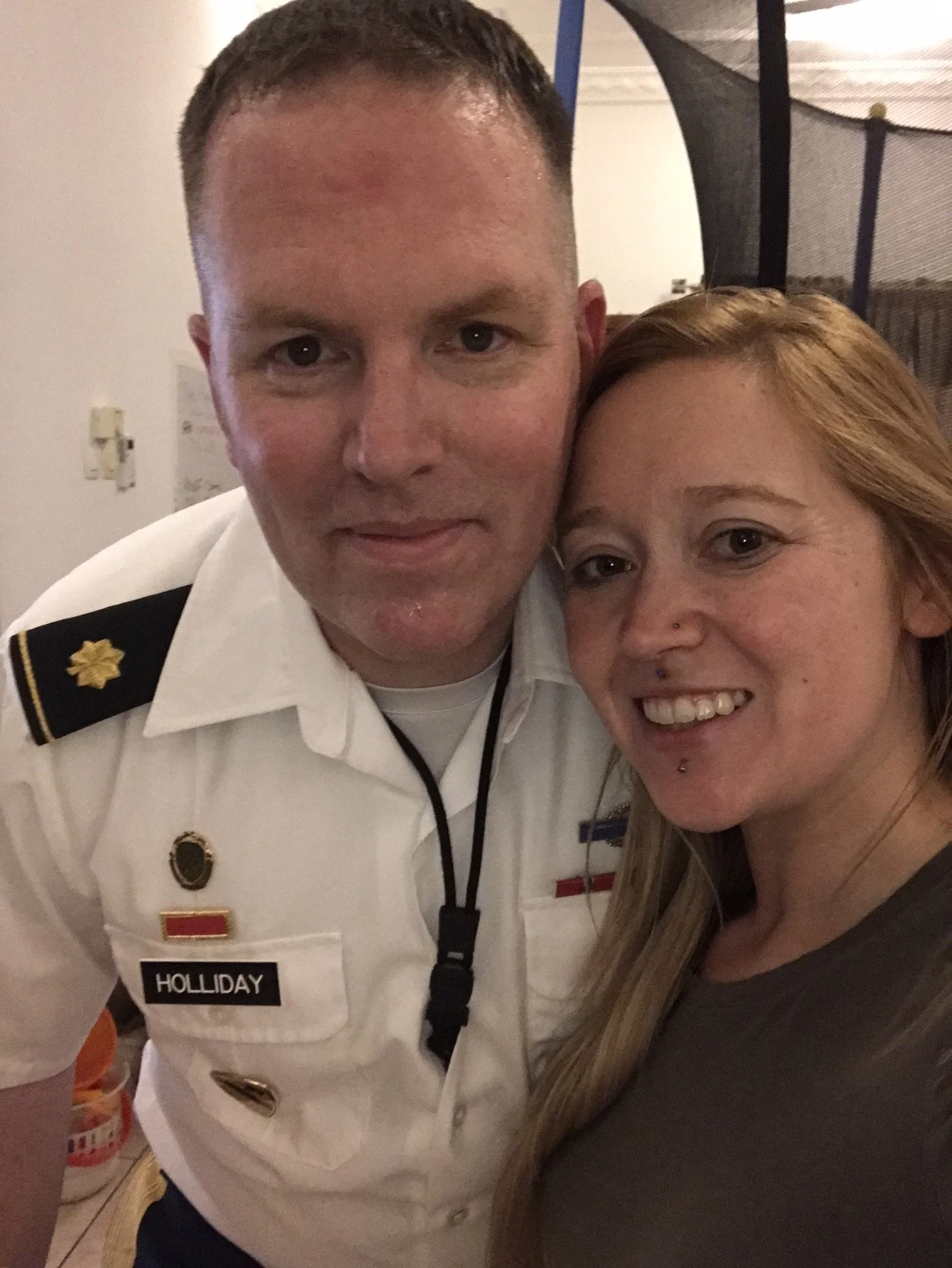 A man and woman taking a close-up selfie together indoors. The man is wearing a white uniform with a name tag reading 'Holliday' and military or service patches on his shoulders. The woman has long, light brown hair and a nose piercing, smiling behin