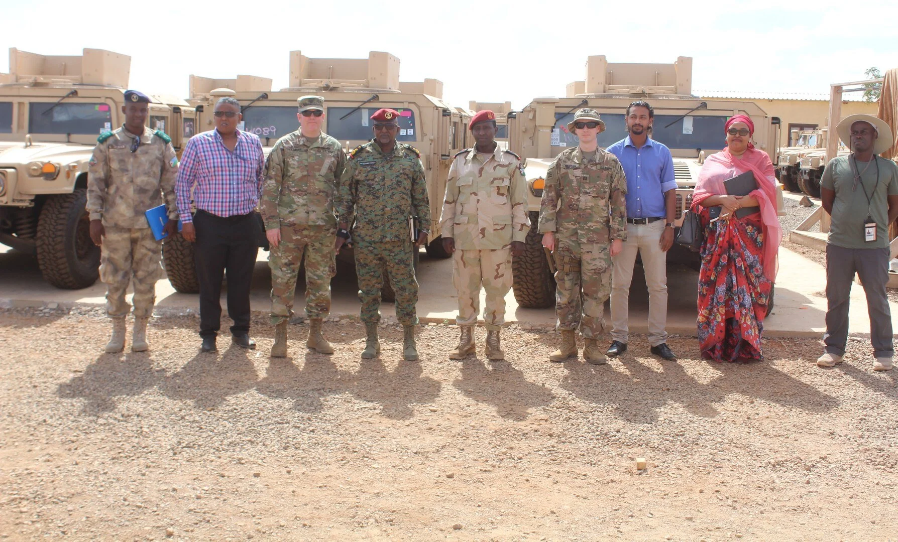 A group of ten people standing in front of military vehicles, including soldiers in camouflage uniforms and civilians in business casual and traditional clothing, outdoors on a sunny day.