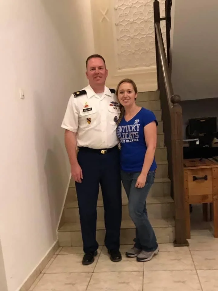 Major Jonathan Holliday in a white military uniform standing next to his wife Jessica in a blue Kentucky Wildcats t-shirt on a staircase indoors.