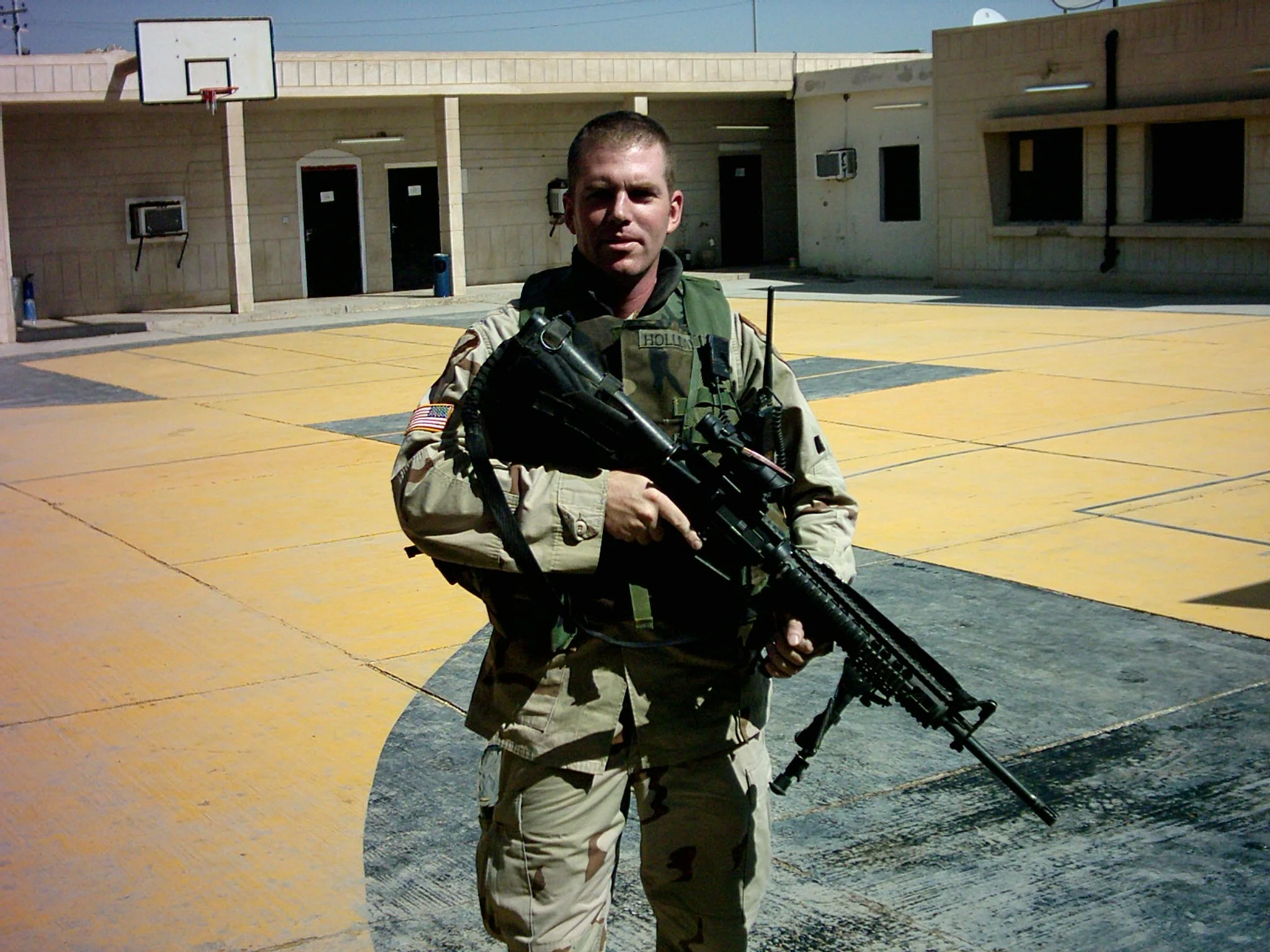 A soldier in camouflage uniform holding a rifle standing in a schoolyard or parking lot with yellow and black painted concrete pavement, a basketball hoop, and a school building in the background.