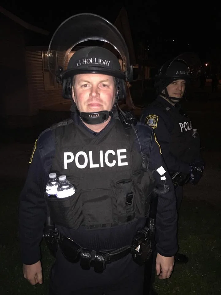 Two police officers in uniform at night. The officer in front is wearing a helmet with a face shield, a black tactical vest with 'POLICE' written on it, and has water bottles in a pouch on his vest. The officer in the background is also wearing a hel