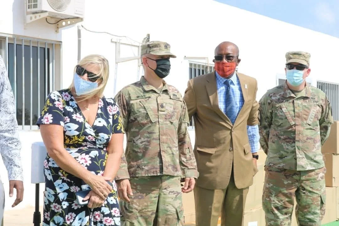 Group of five people, including two in military uniforms and one in a brown suit, standing outdoors with boxes behind them, all wearing face masks.