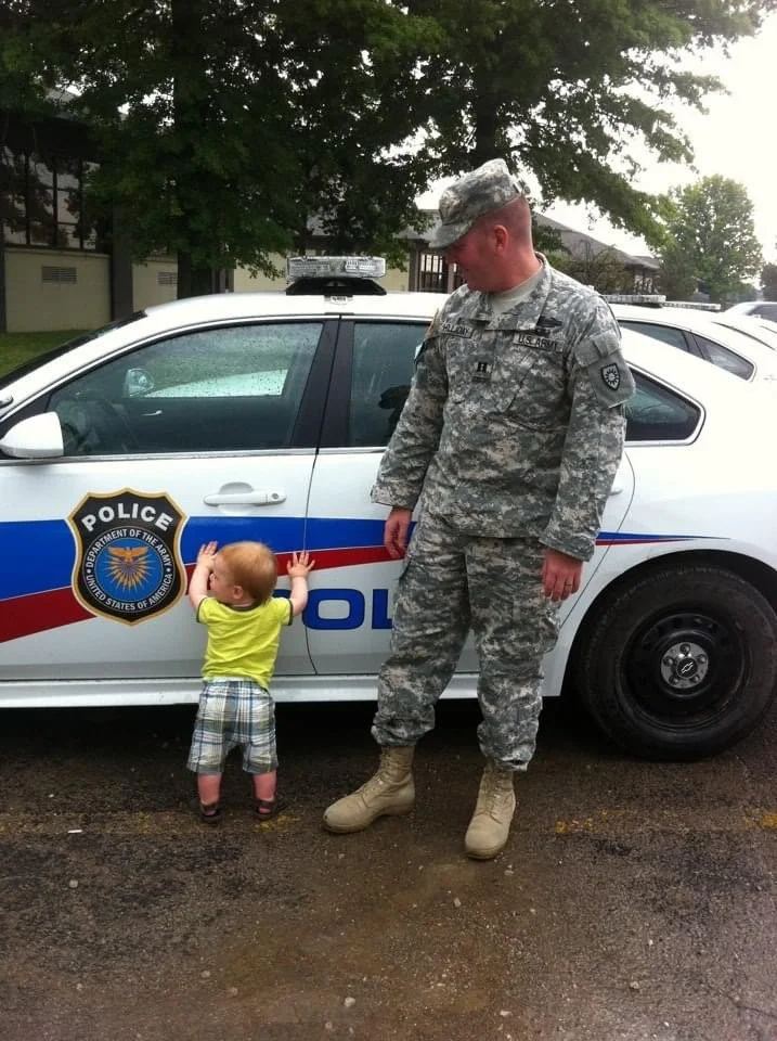 A young child with red hair and a bright yellow shirt pushes against a police car while an adult in military camouflage looks down at the child. The police car is marked with the badge of the United States Department of the Army.