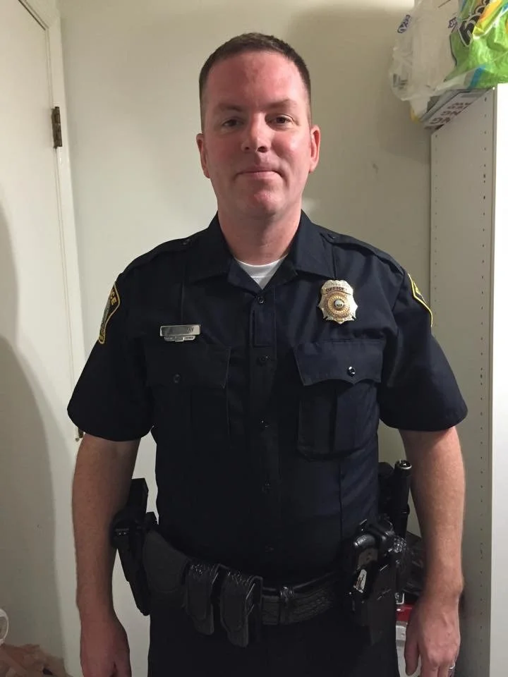 Officer Jonathan Holliday in his Lexington Police uniform standing indoors with a storage shelf in the background.