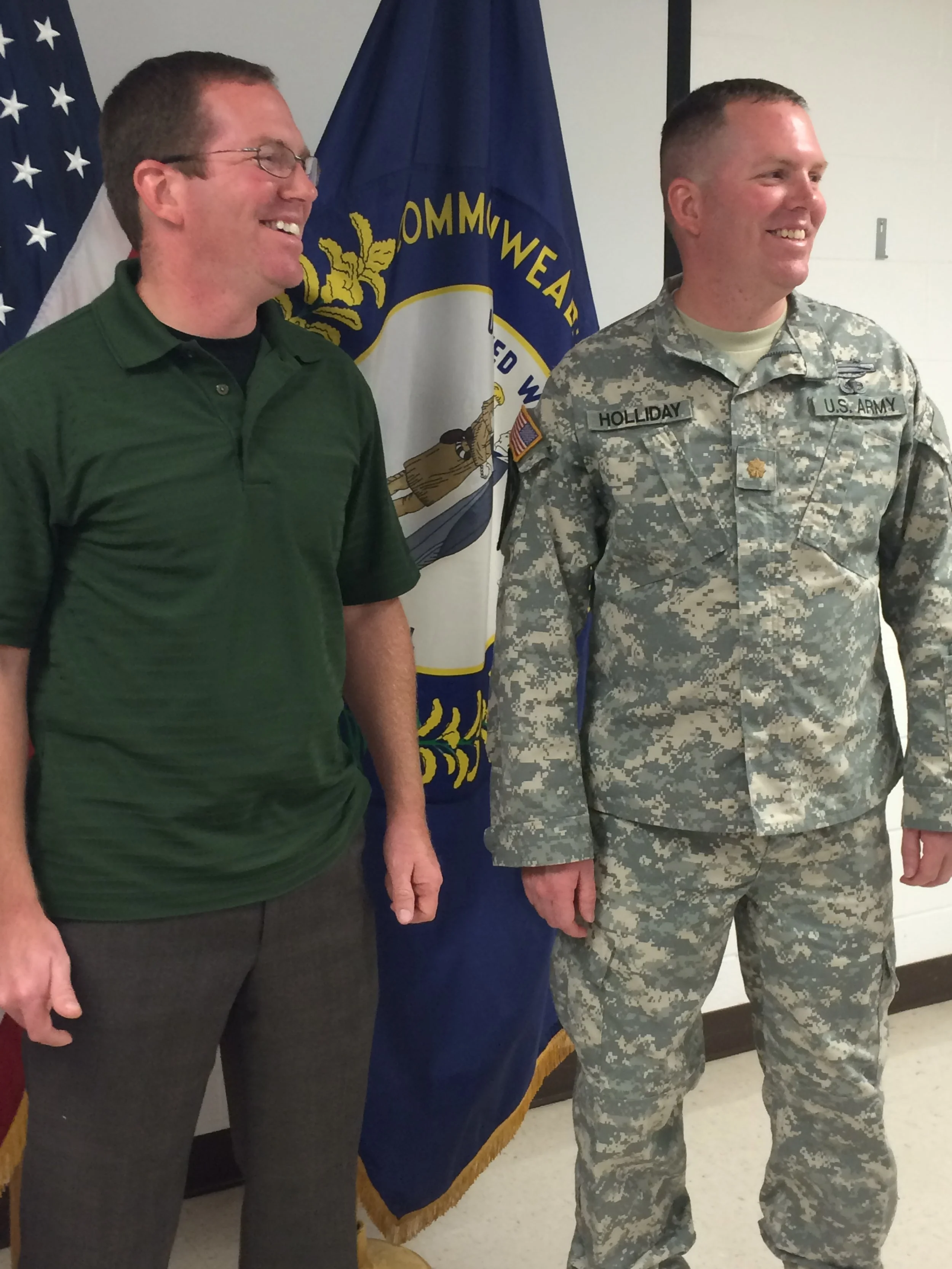 Jonathan Holliday in military uniform and his brother in civilian clothes standing in from of the United States and Kentucky flags, smiling at his promotion ceremony to Major in Richmond, Kentucky.