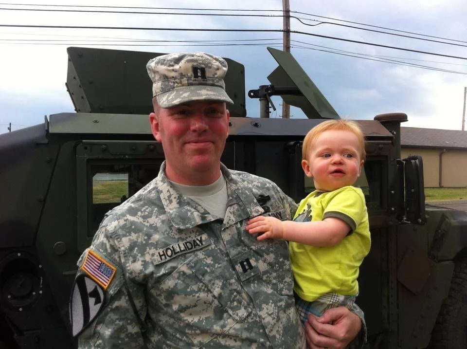 A soldier in uniform holding a young child in front of a military vehicle outdoors.