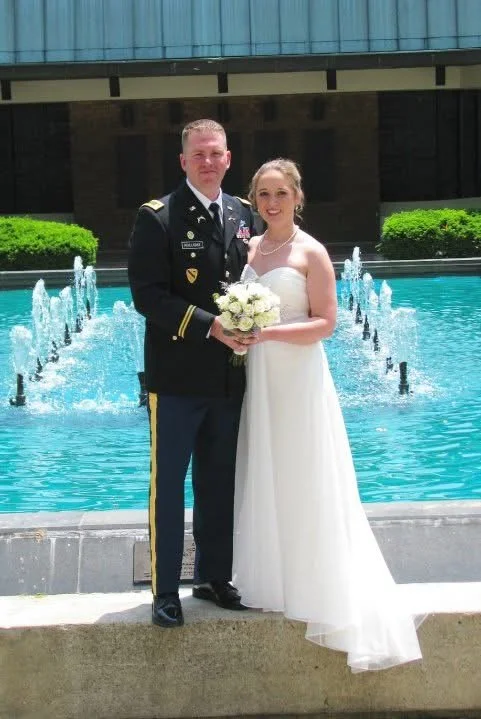 Lieutenant Jonathan Holliday and his wife Jessica at their wedding standing in front of a fountain, the groom in a military uniform and the bride in a wedding dress holding a bouquet of white flowers.