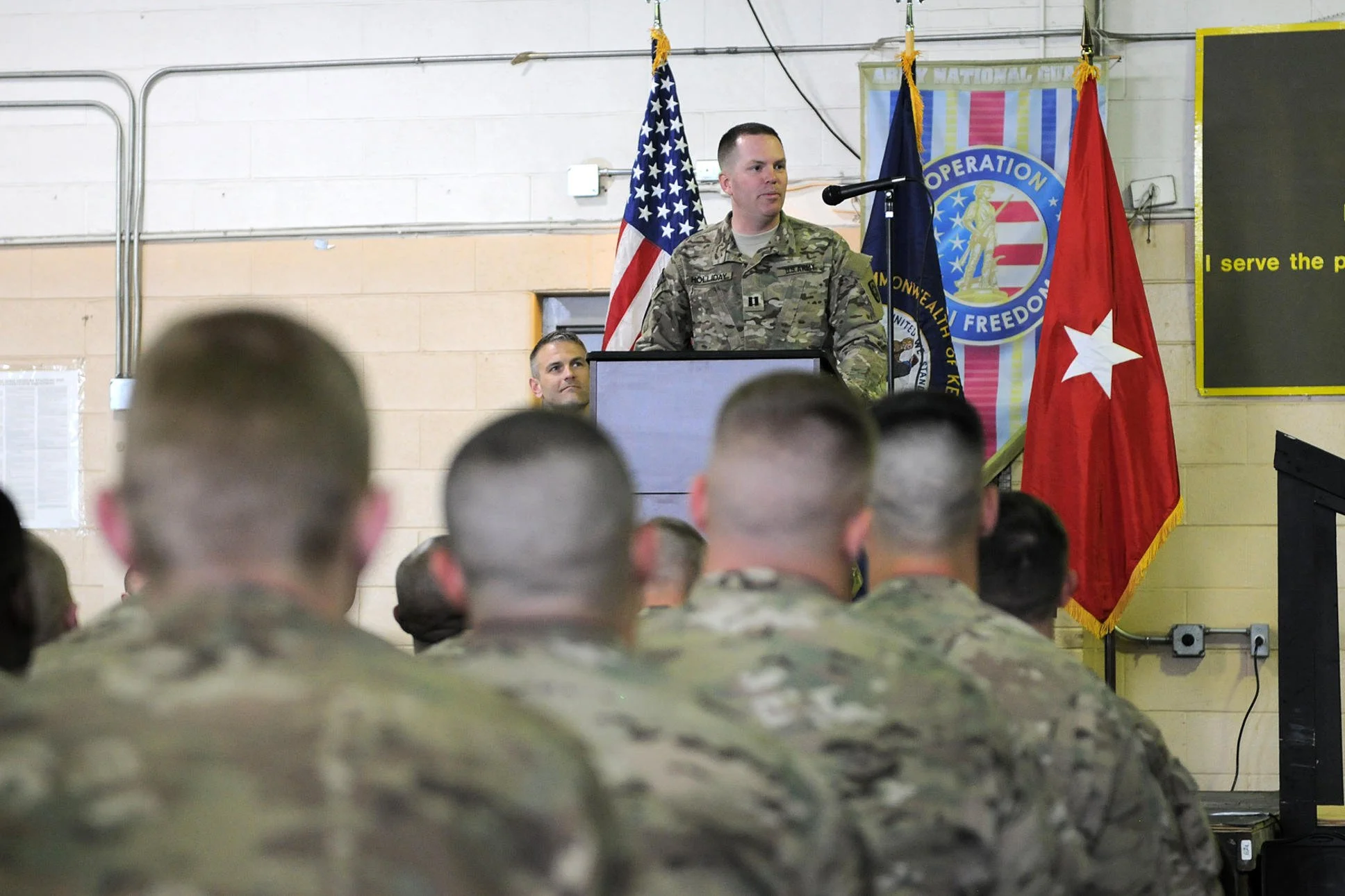 Captain Jonathan Holliday giving a speech at a podium in front of soldiers, with American and other military flags in the background upon his unit's return from Afghanistan.