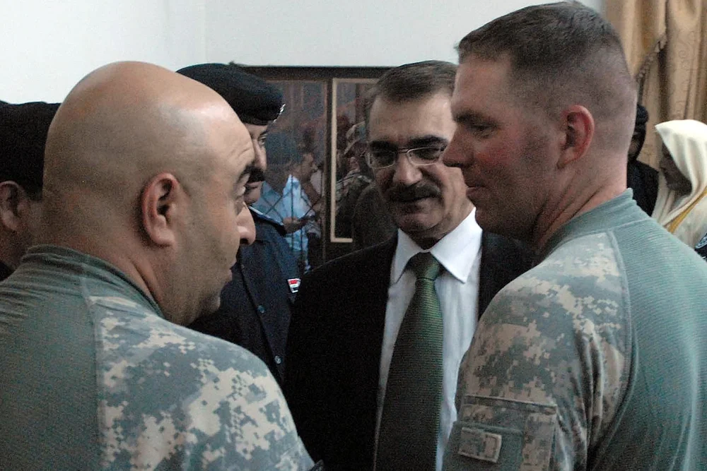 Two military personnel in camouflage uniforms talking to a man in a suit and glasses, in an indoor setting.