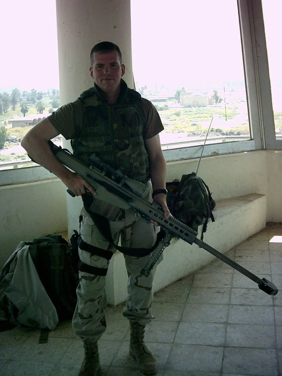 Corporal Jonathan Holliday in military camouflage uniform holding a sniper rifle, standing indoors near large windows showing an outdoor view in Baghdad, Iraq.