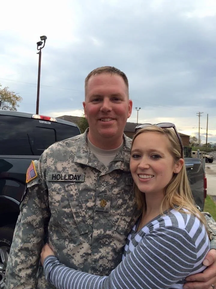 A smiling man in military uniform embracing a woman in a striped shirt outdoors on a cloudy day.