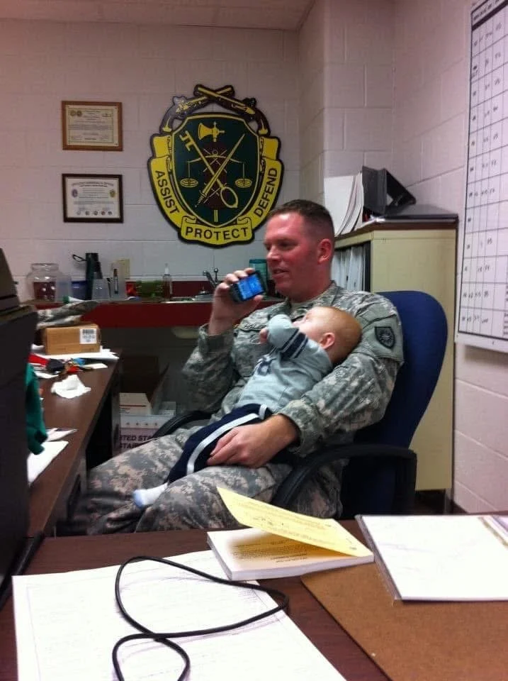 A man in military camouflage uniform sitting at a cluttered desk, talking on a cell phone while holding a sleeping young child in his lap. The office has framed certificates on the wall and a shield-shaped emblem with the words 'Protect, Assist, Defend' in the background.
