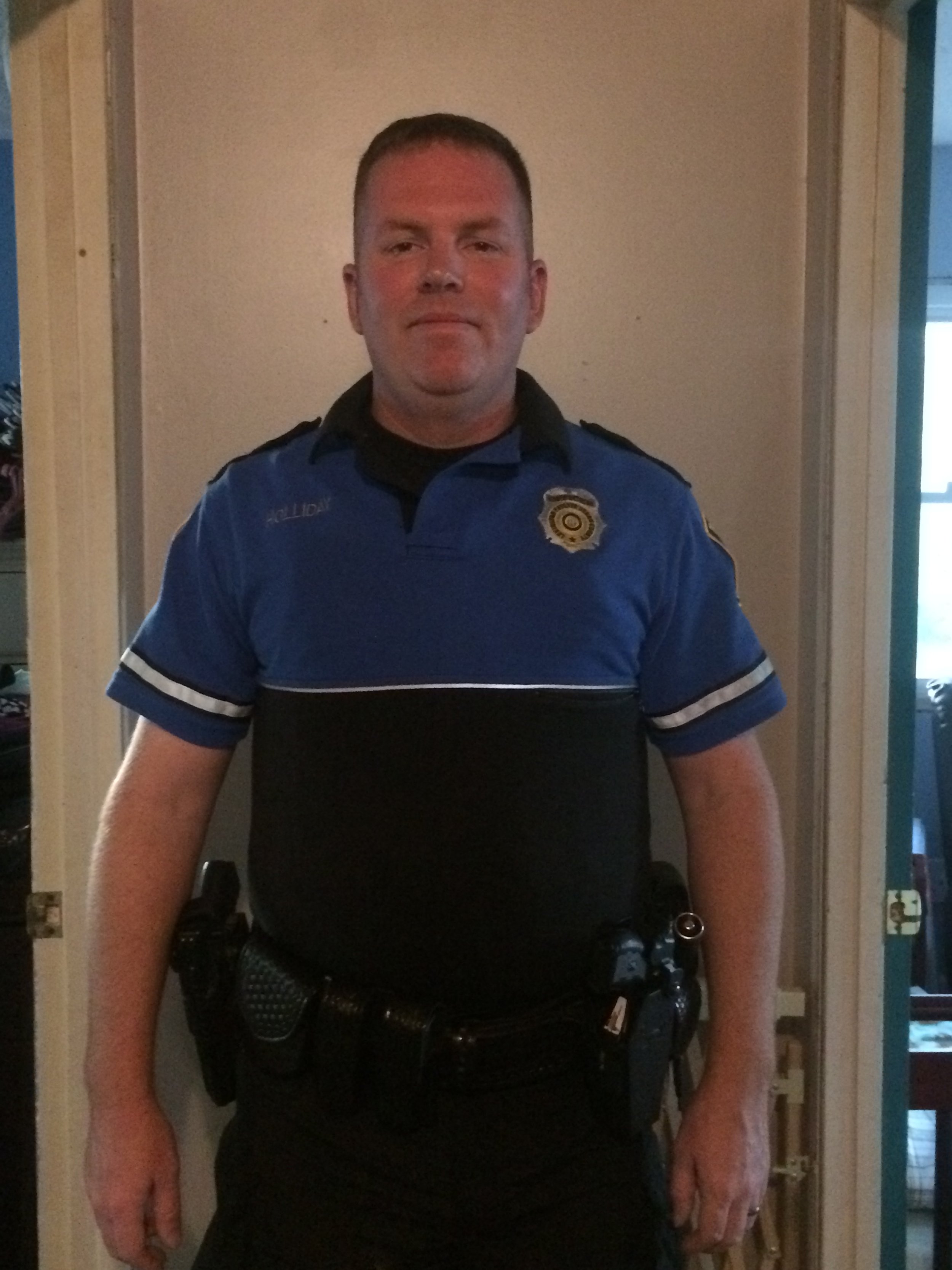 A male police officer wearing a blue uniform with a badge, standing indoors against a beige wall.