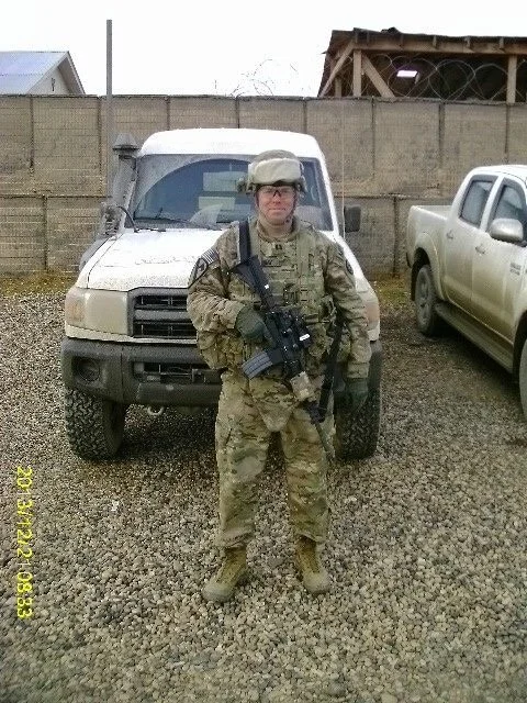 Captain Jonathan Holliday in camouflage uniform standing in front of a beige military vehicle, holding a rifle, with a pickup truck to the right, and a wooden structure and fence in the background in Northern Afghanistan.