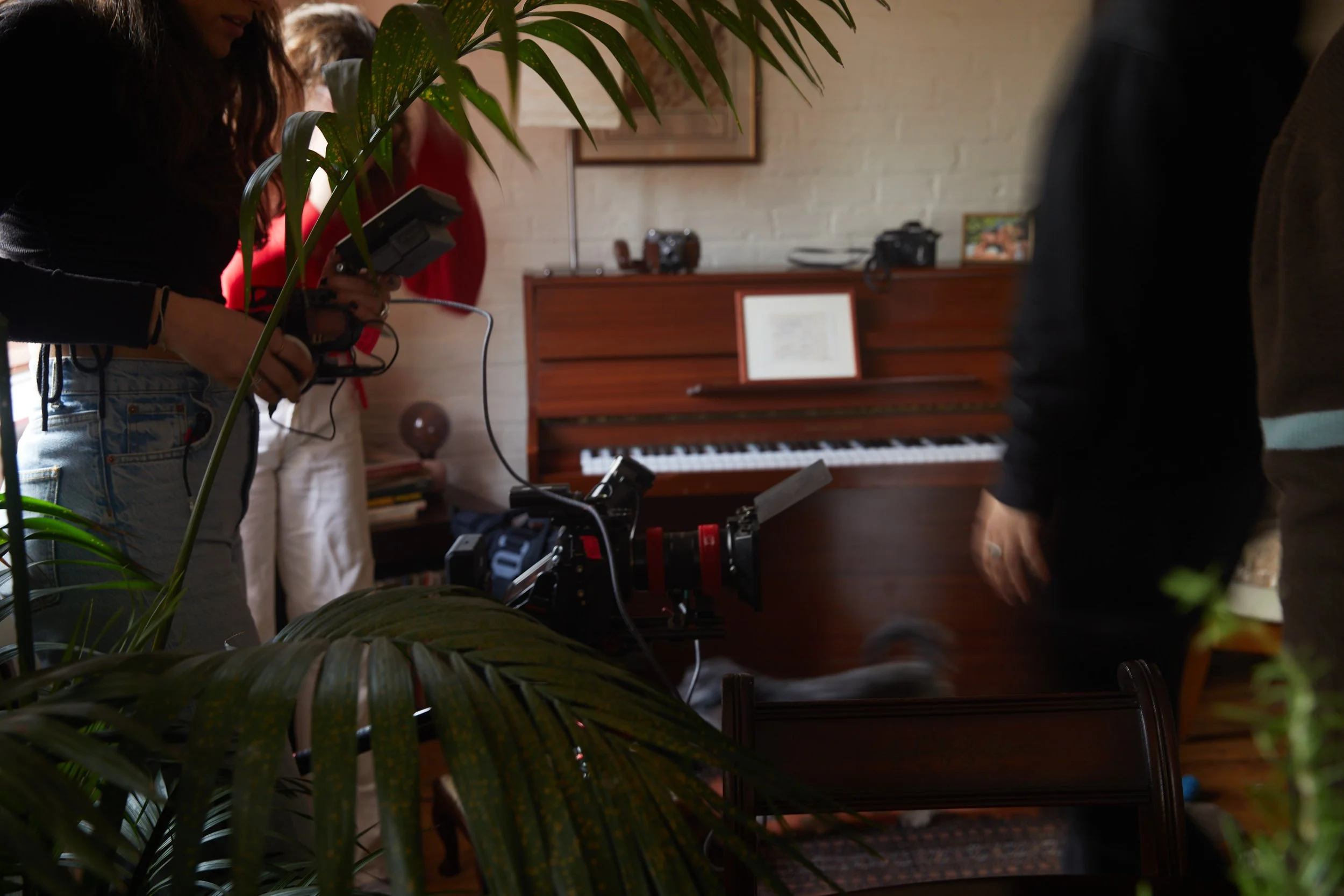 A film crew setting up a scene in a music room with a piano, cameras, and crew members.