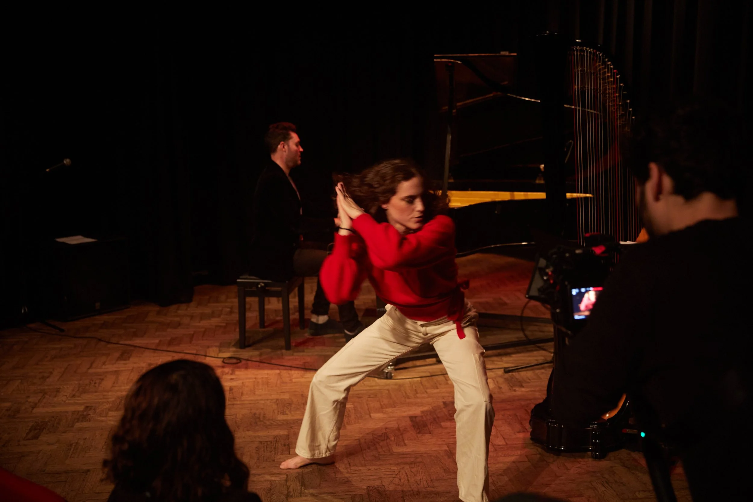 A woman wearing a red top and beige pants dancing on stage with a pianist in the background and a camera operator filming her.