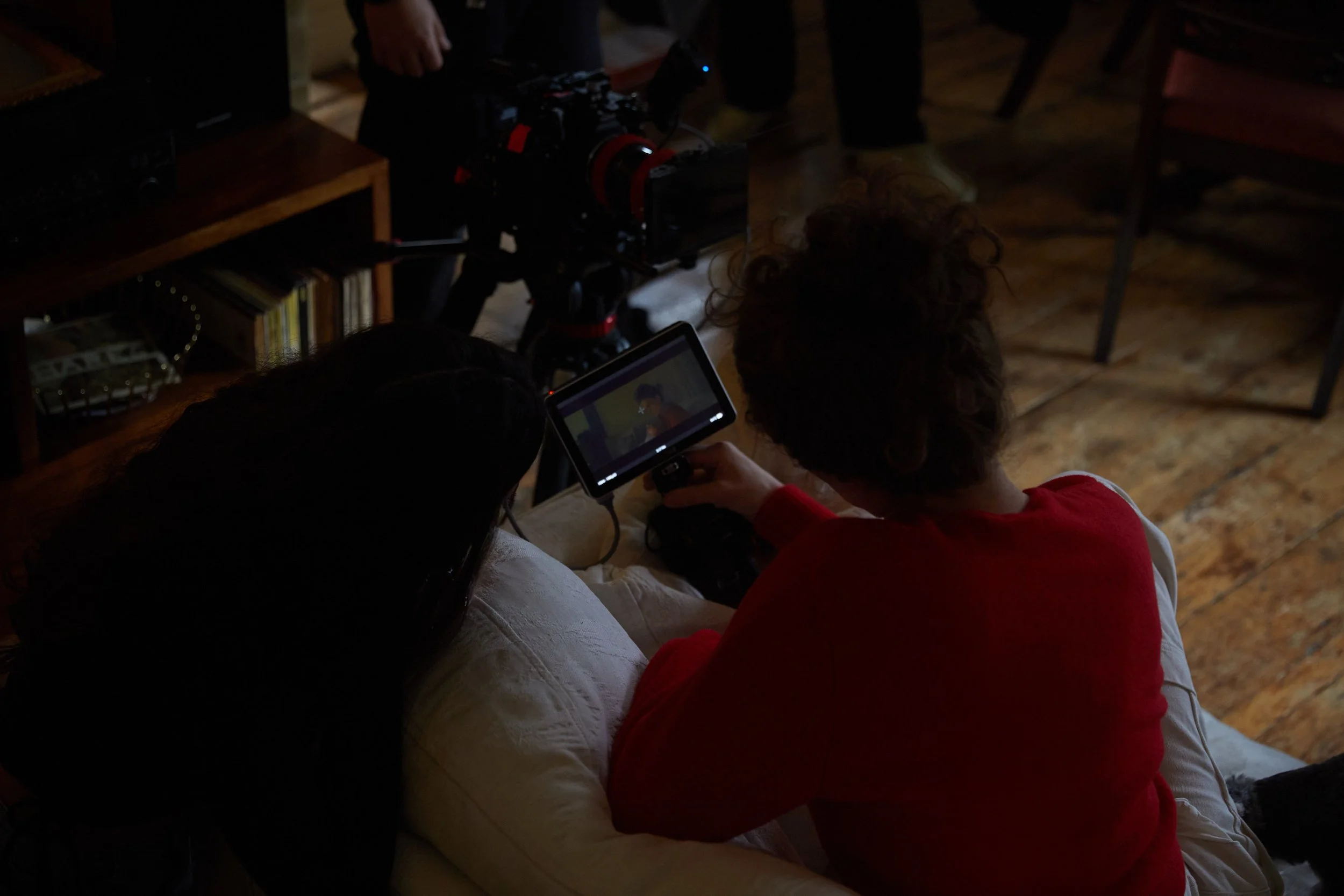 Two people seated on a couch watching a small video monitor, with filming equipment and a camera on a tripod nearby in a dimly lit room.