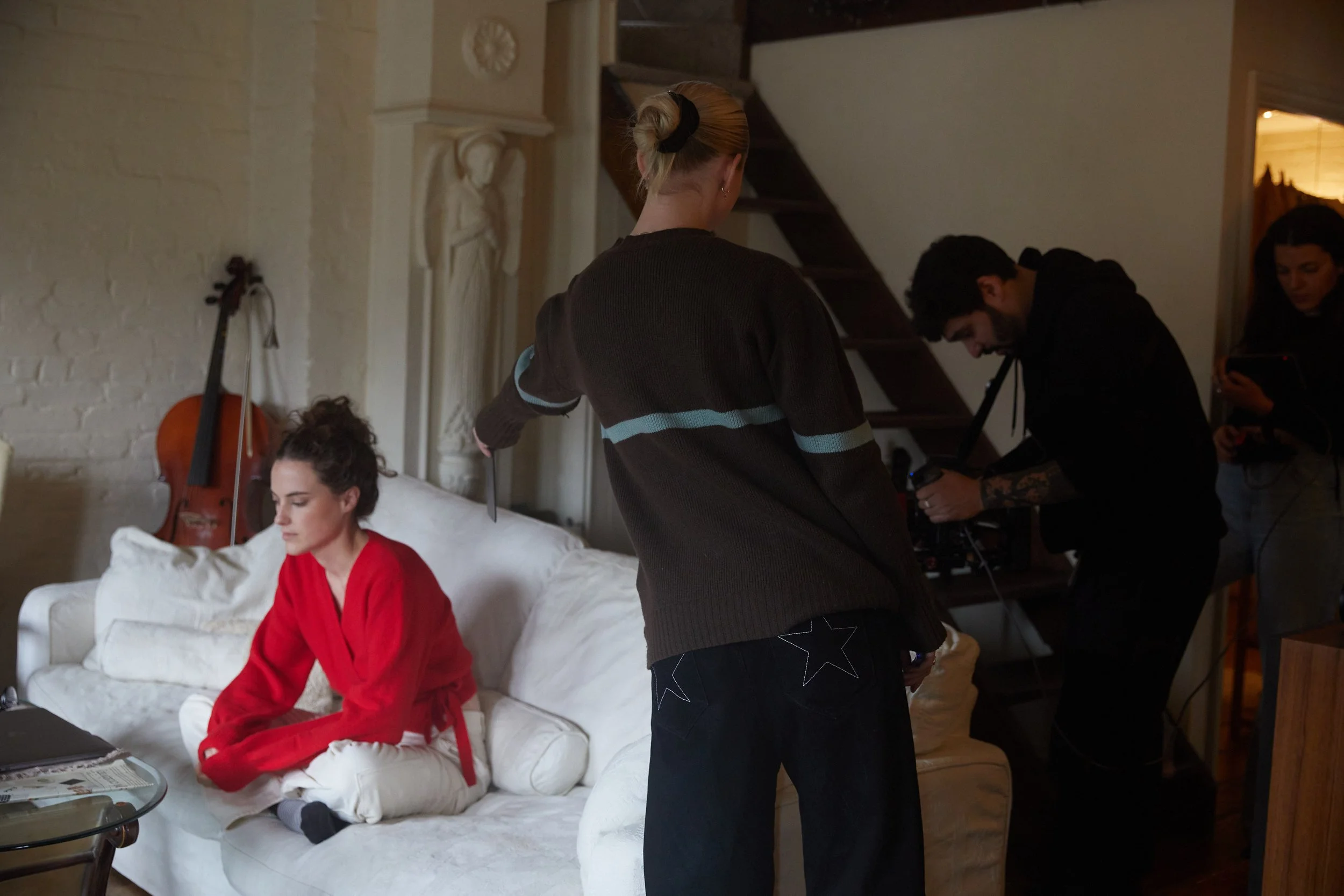 A woman wearing a red robe sits on a white couch during a photo shoot or filming. A crew member with a ponytail and sweater is holding a reflector or diffuser near her. Two other crew members are operating camera and equipment nearby. The setting is