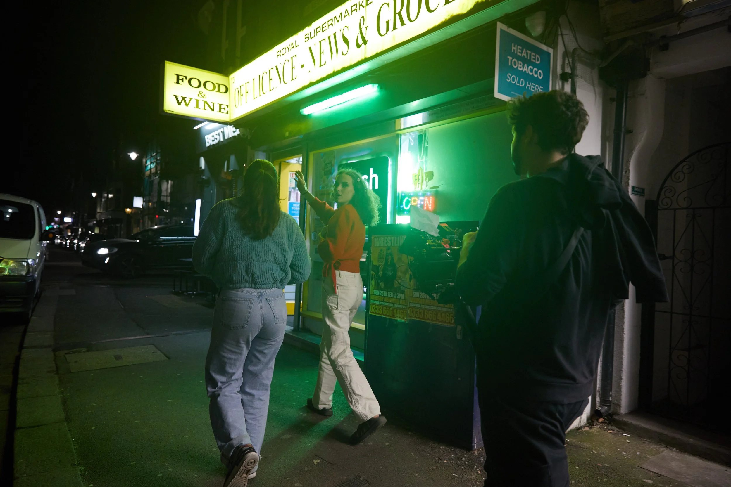Nighttime street scene outside a convenience store with three people near the entrance, a woman posing and two others walking or filming. The store has neon and illuminated signs, including one for 'Food & Wine' and 'Office, News & Groceries'.