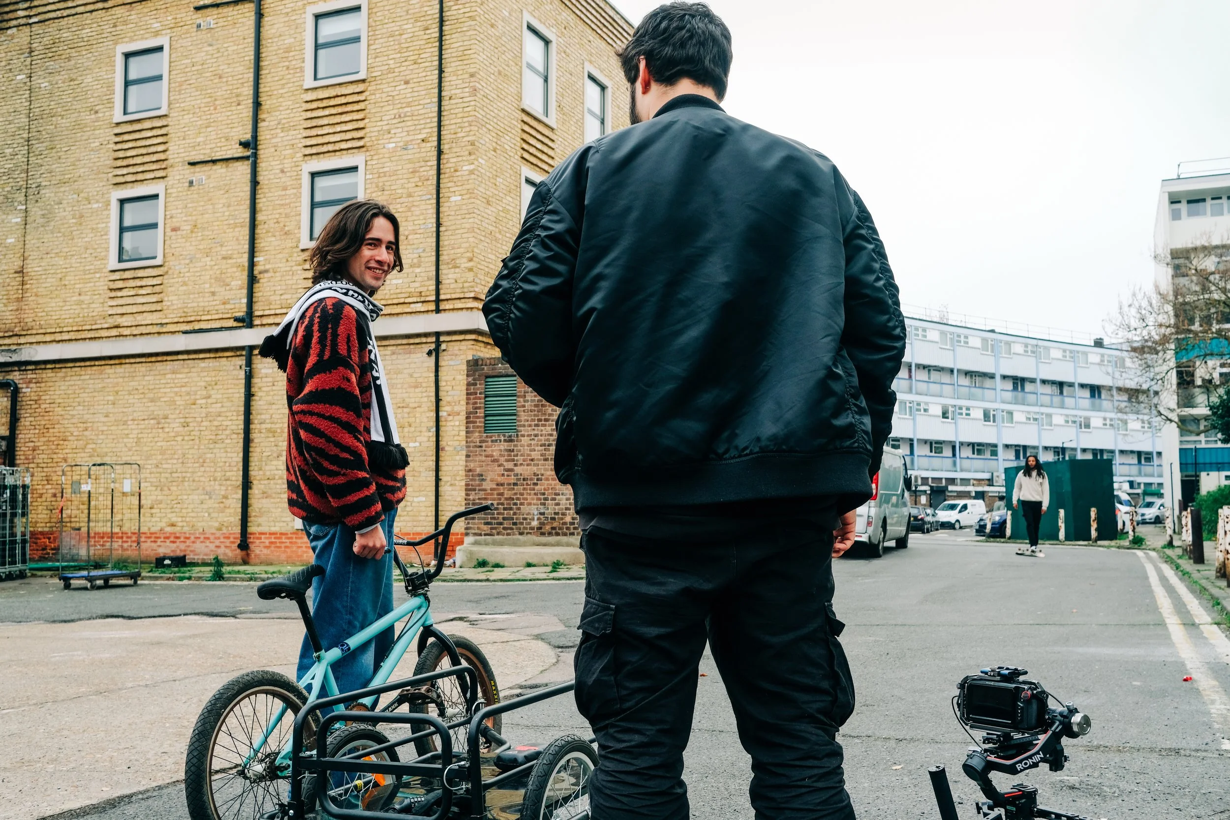 A young woman with curly hair, wearing a red and black patterned sweater and a scarf, stands next to a blue bicycle and is smiling at a man with dark hair, wearing a black jacket. They are outside near a brick building with other people and parked ca