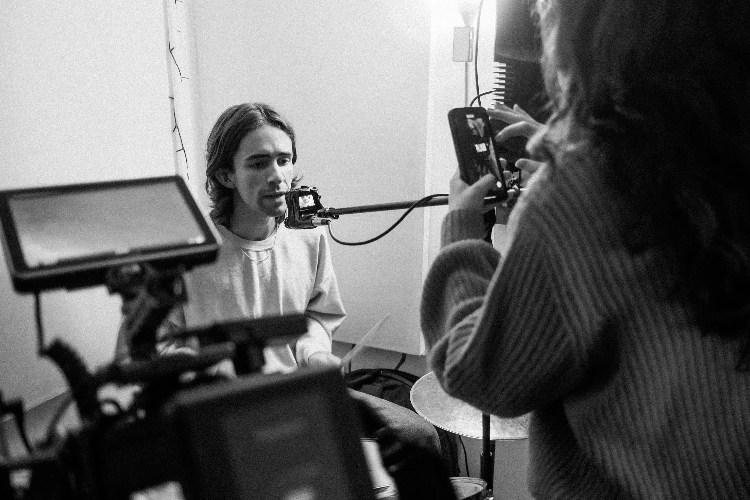 A person with long hair, wearing a striped sweater, is filming or photographing a young man with shoulder-length hair who is sitting in front of a microphone, in what appears to be a recording or interview setup in a small room.