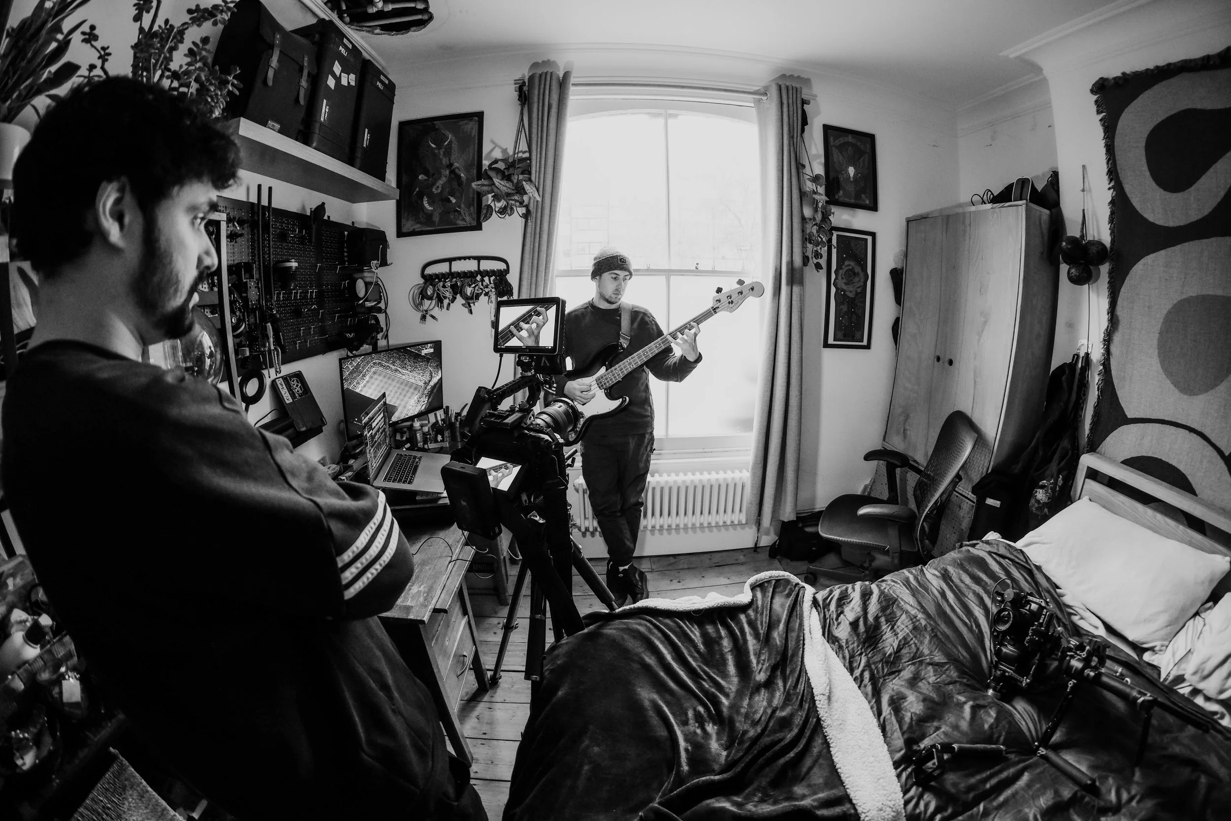 A person is playing a guitar in a bedroom while being filmed by another person with a camera. The room has a bed, a wardrobe, and various decorations and equipment.