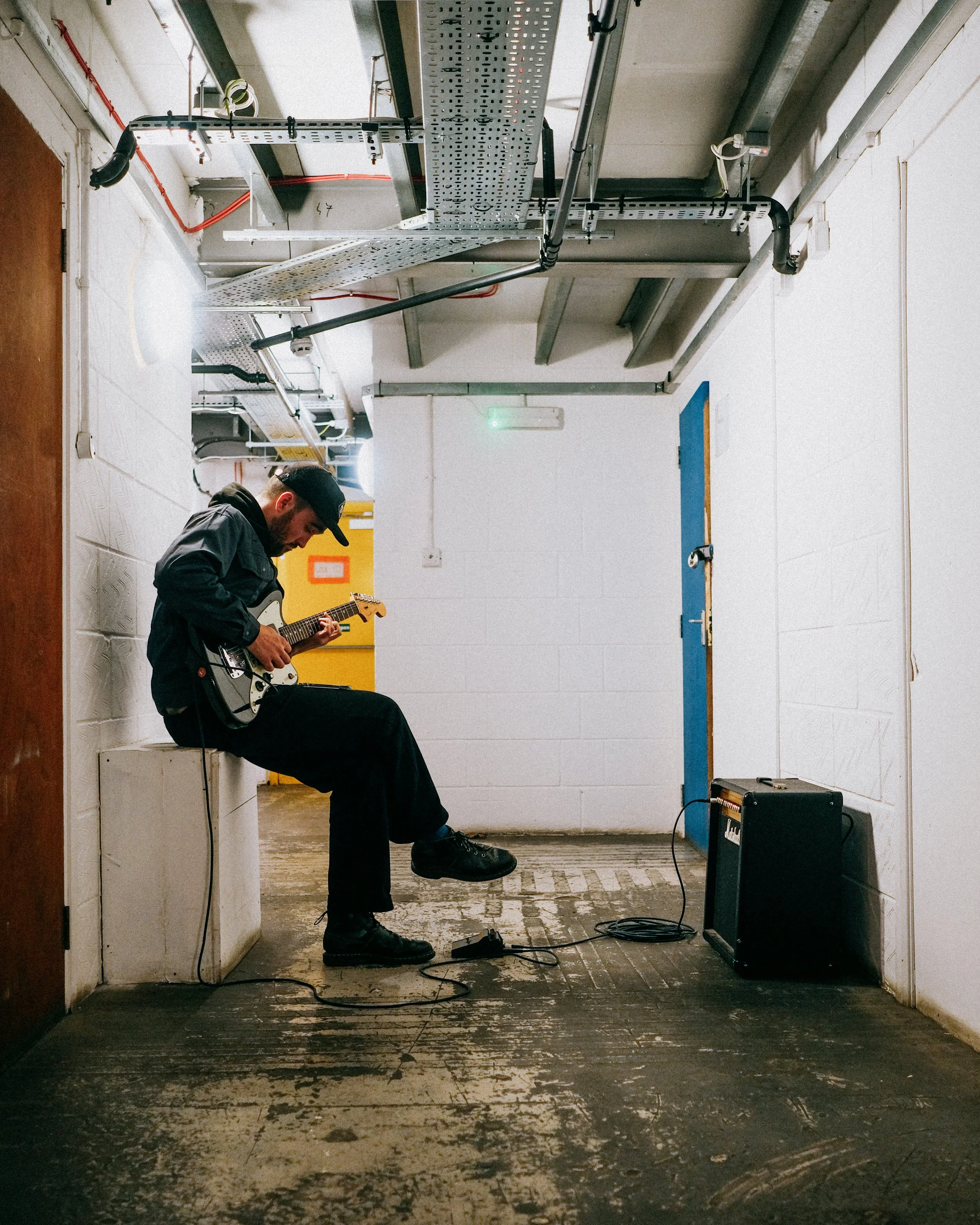 A man wearing a black jacket, black pants, and a black cap sits on a white block in a basement or industrial space, playing an electric guitar. An amplifier is on the floor to his right, with a cable connected to his guitar. The ceiling has exposed p