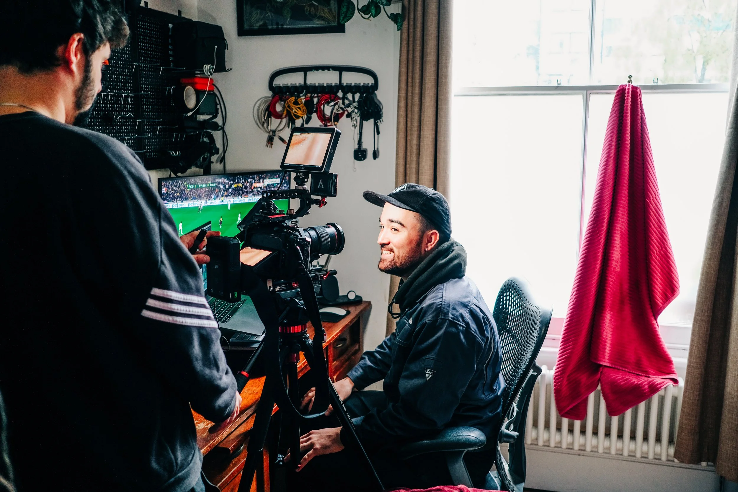 A man sitting in an office chair is being filmed while watching a sports game on a monitor. Another person operates the camera in front of him. The room has a window with beige curtains and a red blanket hanging on a radiator.