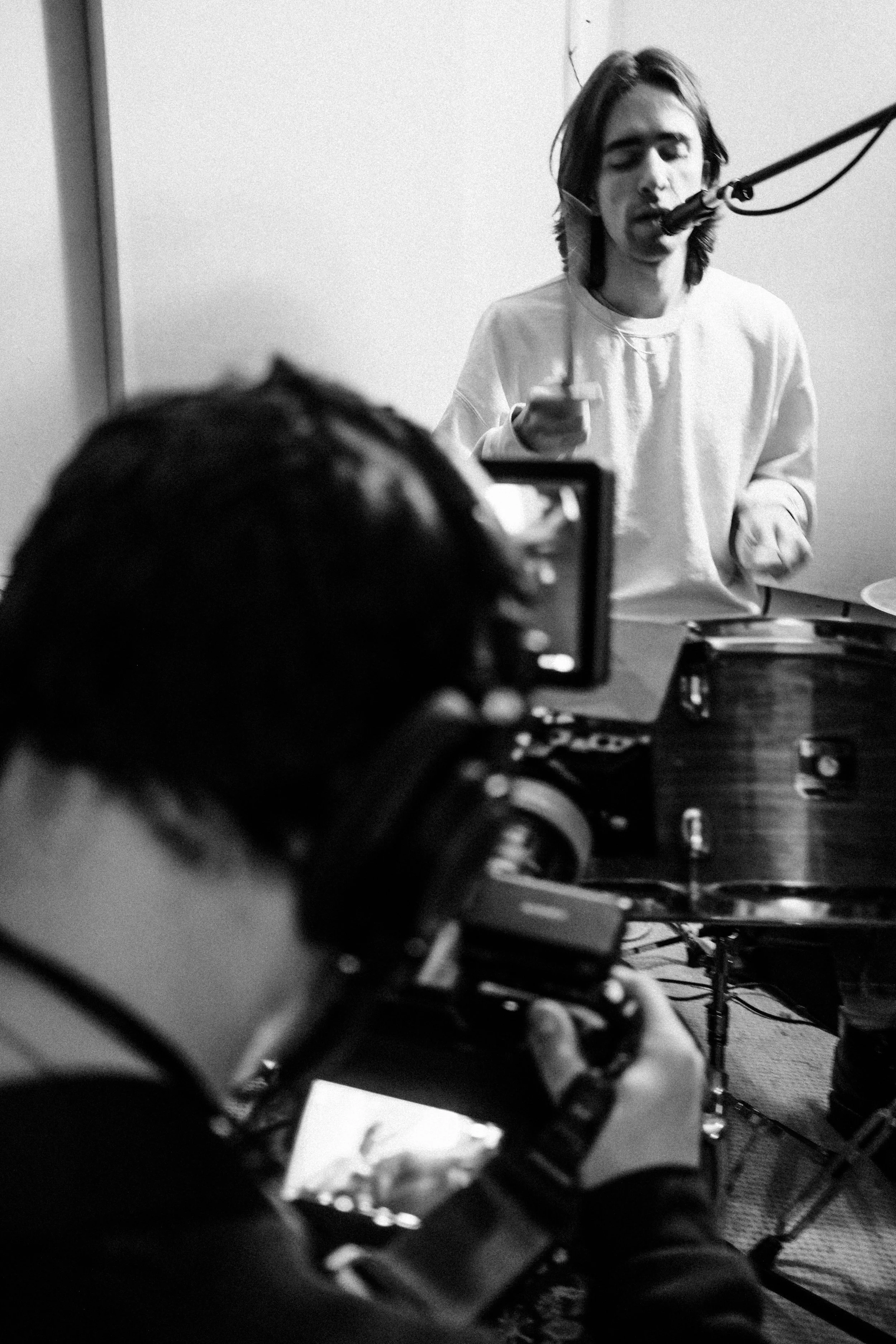 A person is recording a musician singing into a microphone while playing drums in a studio.